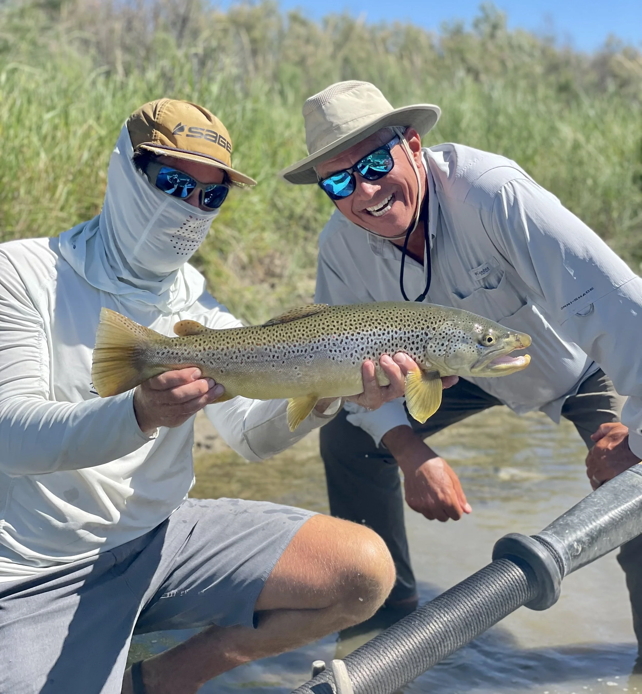 Two men wearing hats, sunglasses, and white shirts holding a large rainbow trout while standing in a river.