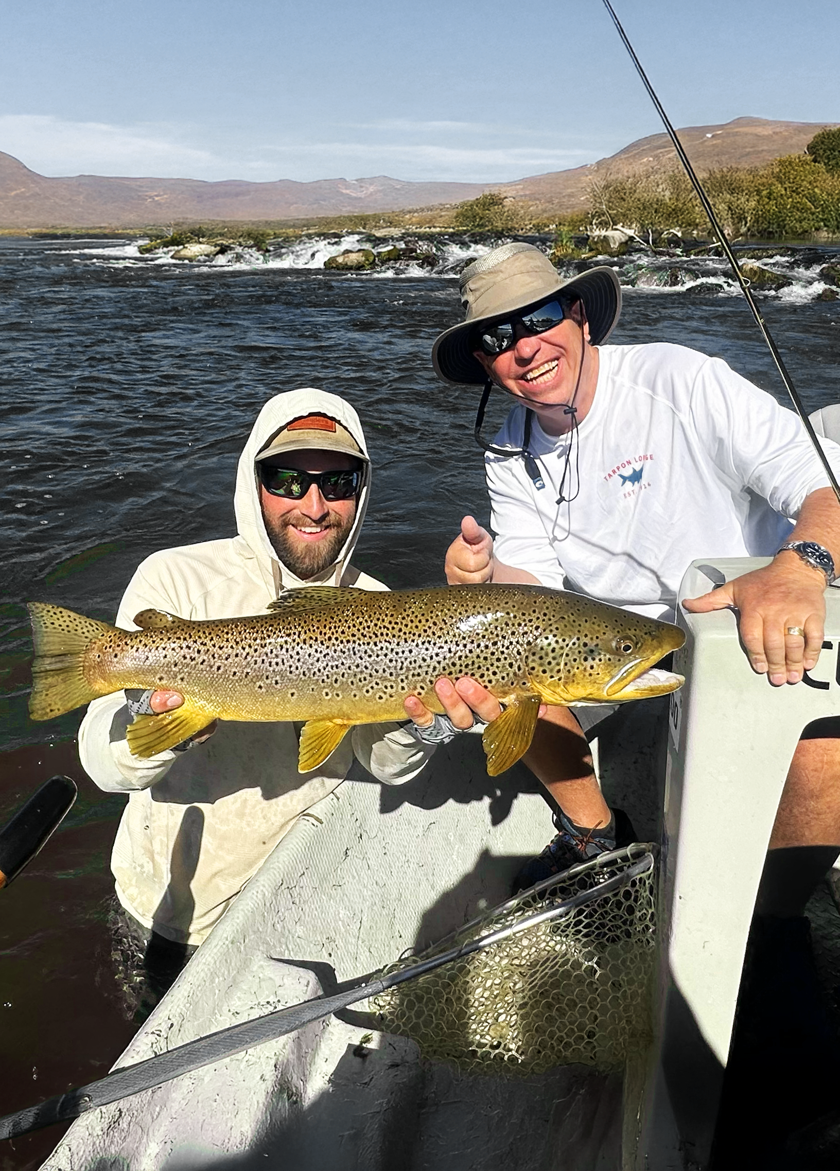 Two smiling men on a boat in a river holding a large rainbow trout.