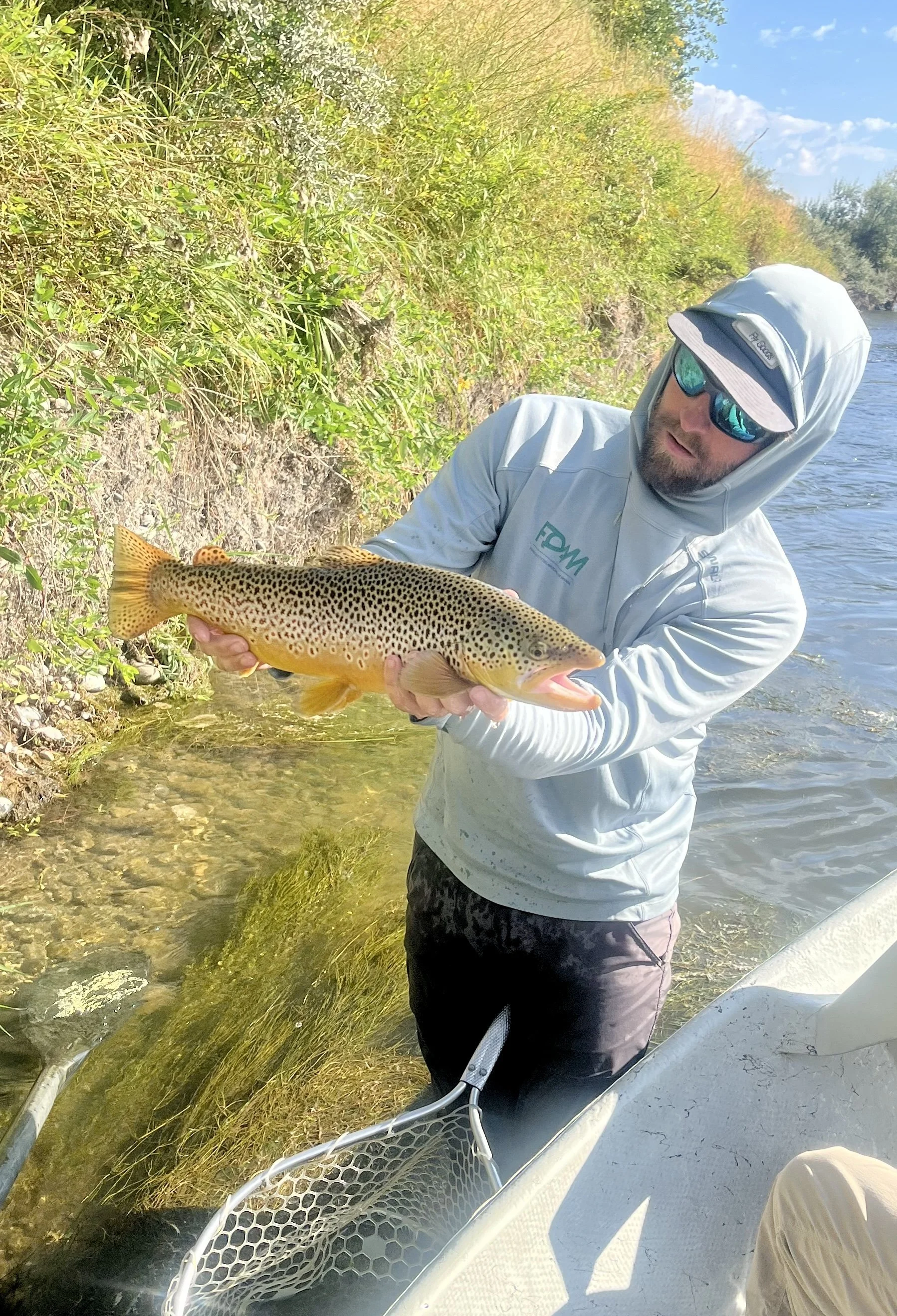 A man wearing sunglasses, a hoodie, and a fishing shirt holding a large fish near a riverbank.