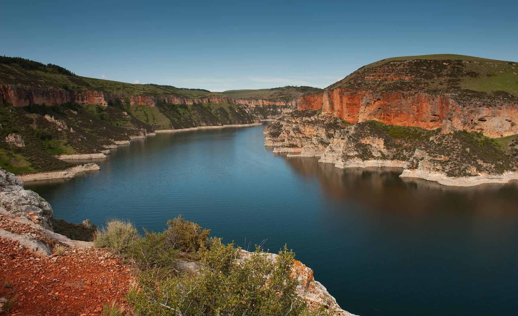 A wide view of a river flowing between steep red rock cliffs with patches of green vegetation under a clear blue sky.