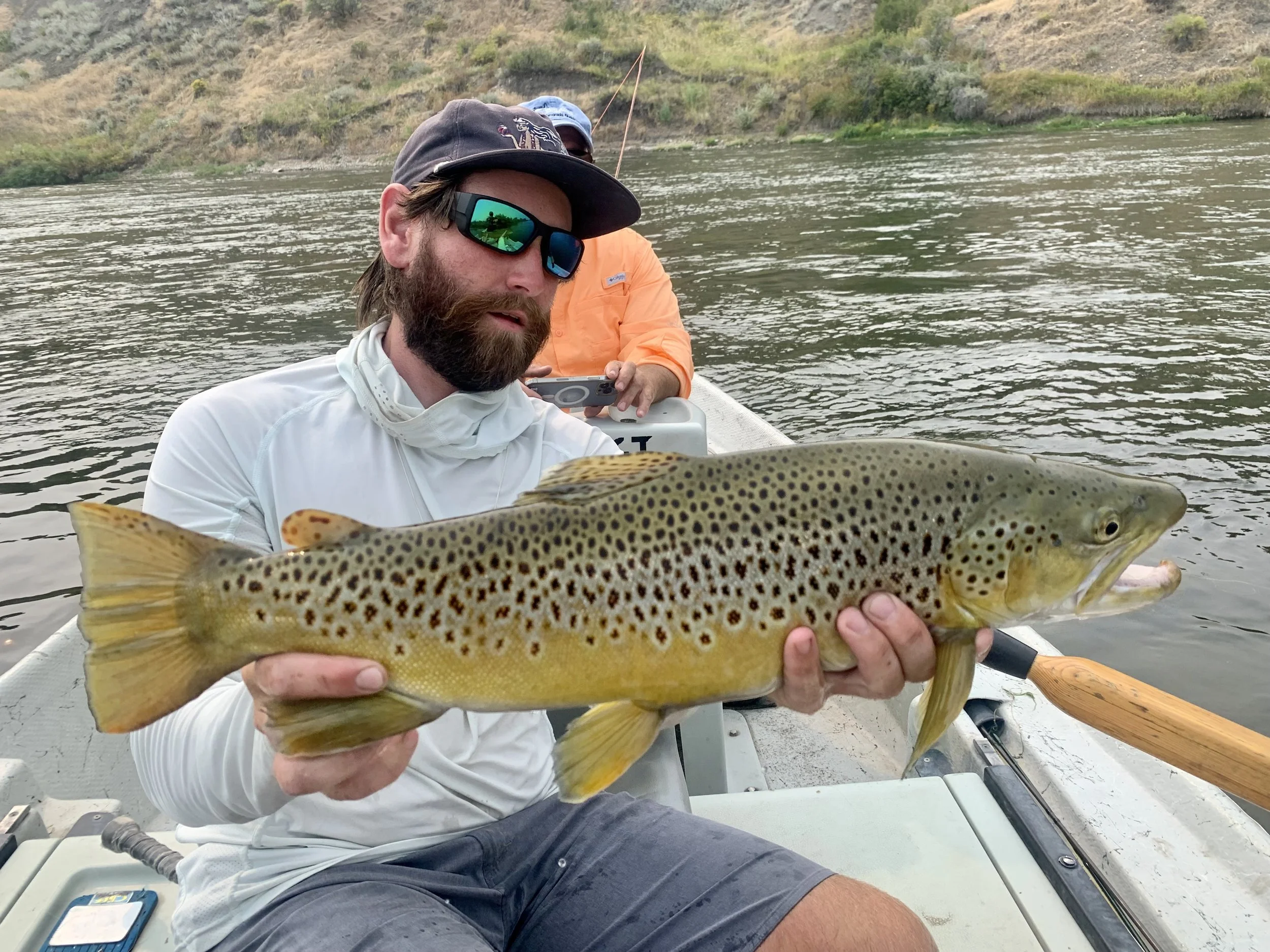 Man with a beard wearing sunglasses holding a large trout fish on a boat in a river, with another person fishing in the background.