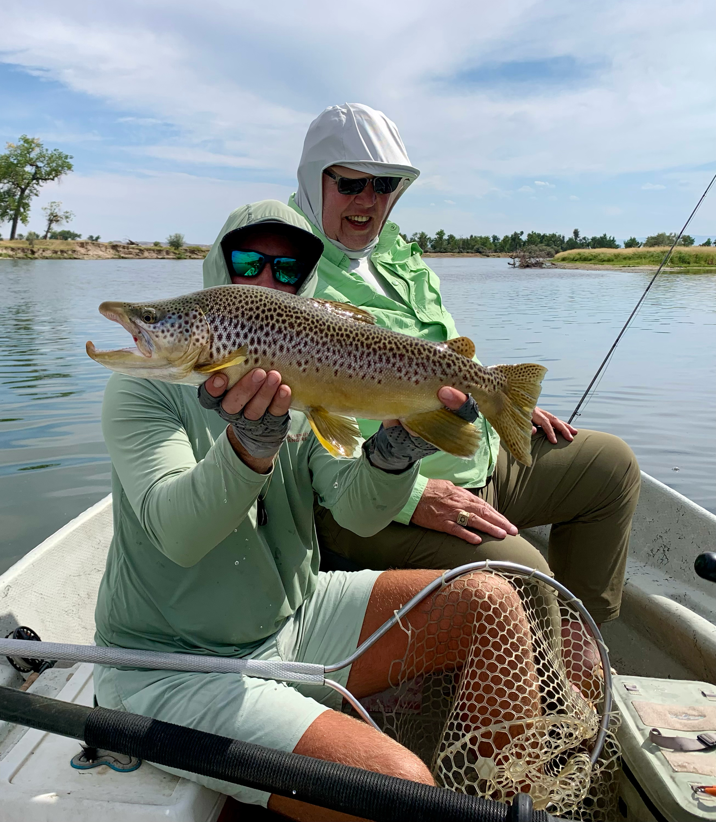 Two people in a boat holding a large rainbow trout with a scenic lake and shoreline in the background.