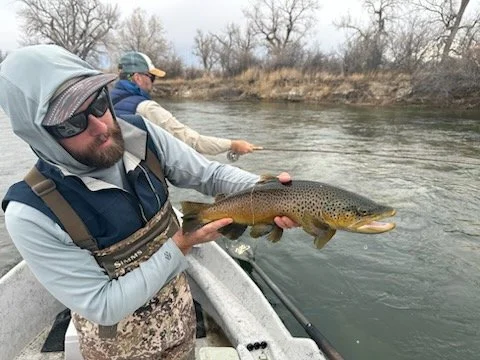 Man wearing sunglasses and a hooded jacket holding a large fish while on a boat, with another man fishing in the background.