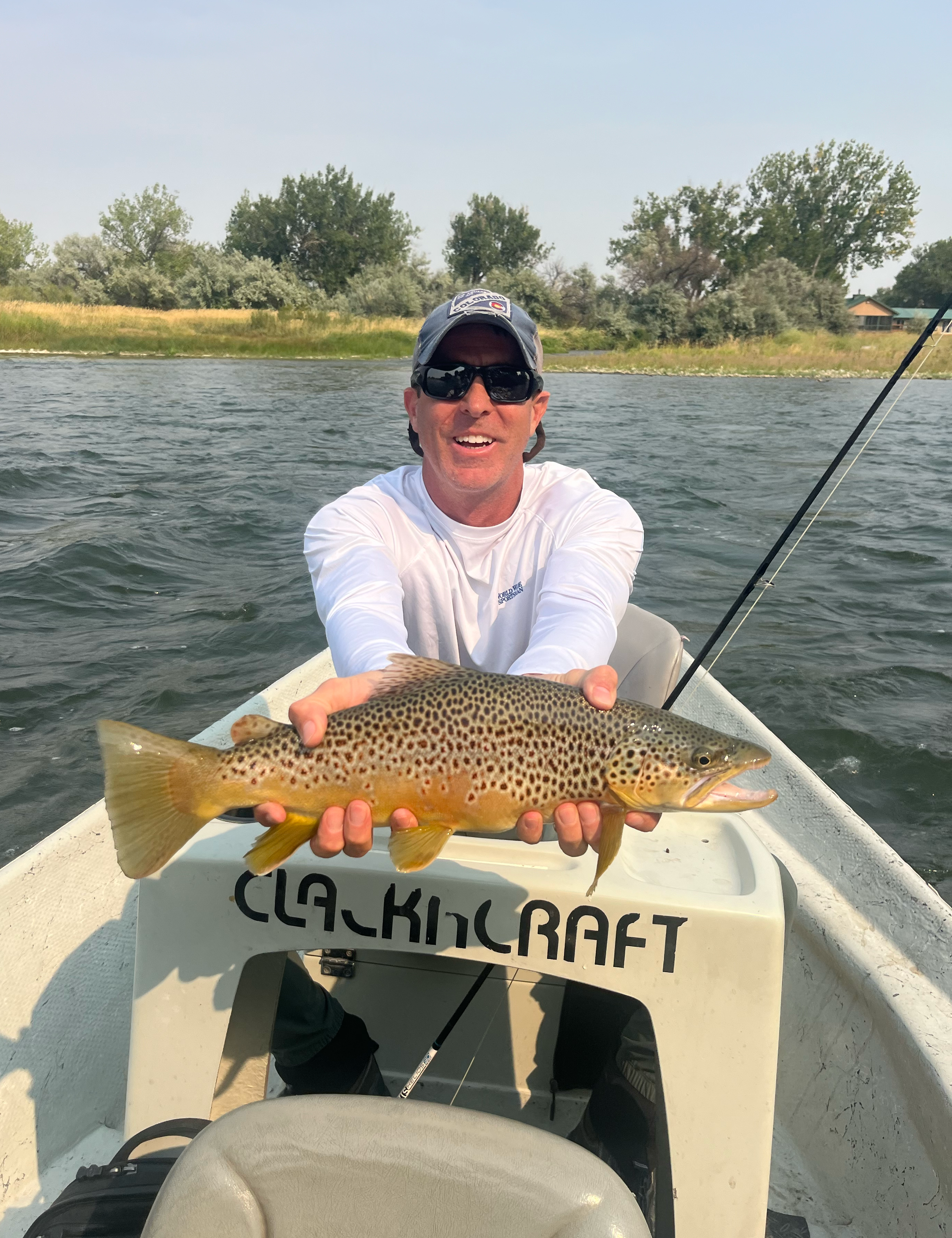 A man in sunglasses and a white shirt on a boat holding a large spotted fish he caught, with a fishing rod in the background and water and trees behind him.