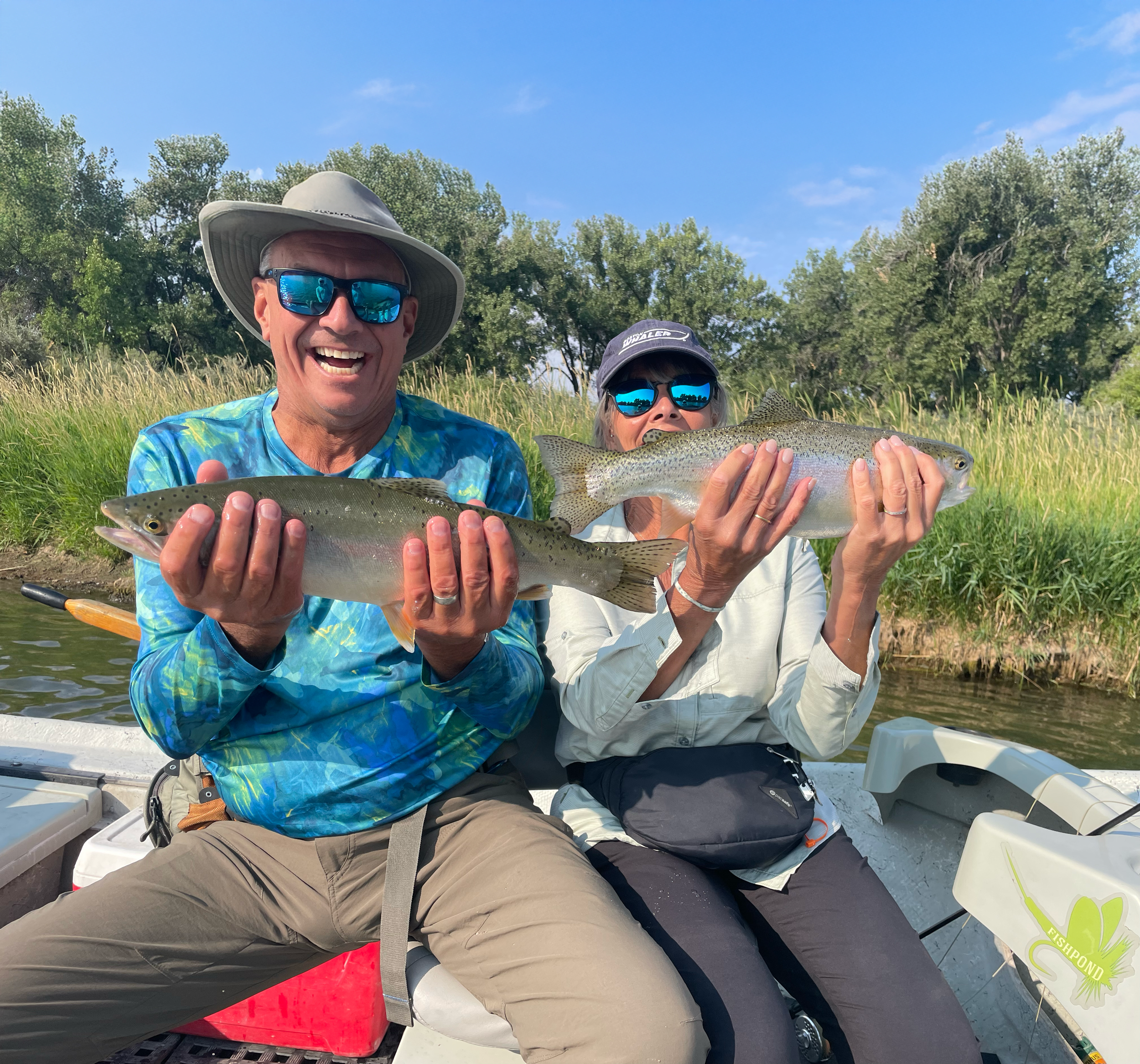 A smiling man and woman holding large fish while sitting in a boat on the water, with trees and grass in the background.