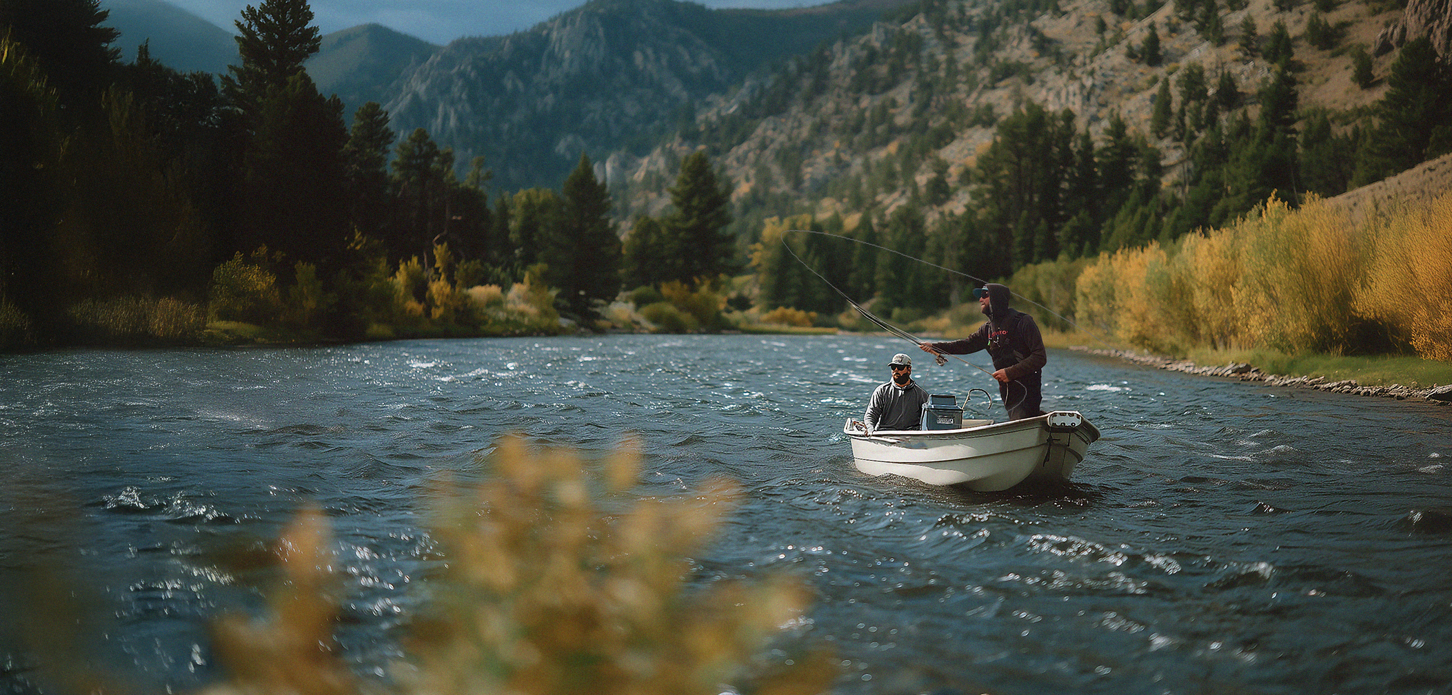 Two men in a boat fishing on a river surrounded by trees and mountains with overcast sky.