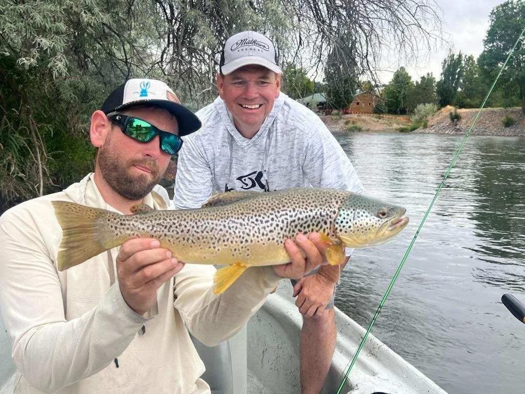 Two men on a boat by a river, one of them holding a large fish, likely a trout, with trees and a house in the background.