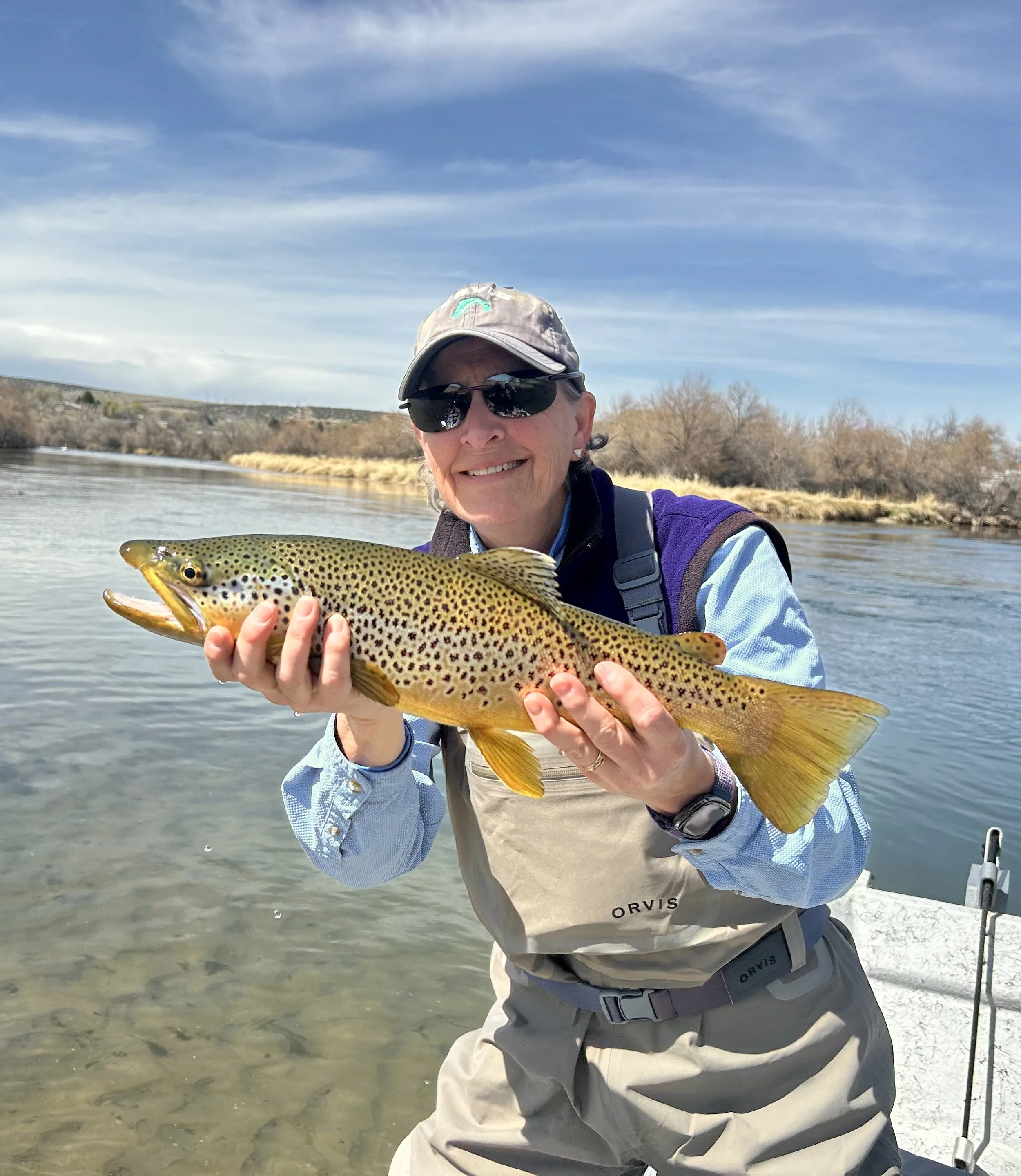 A woman wearing sunglasses, a hat, and outdoor clothing holding a large rainbow trout fish near a river, smiling at the camera.