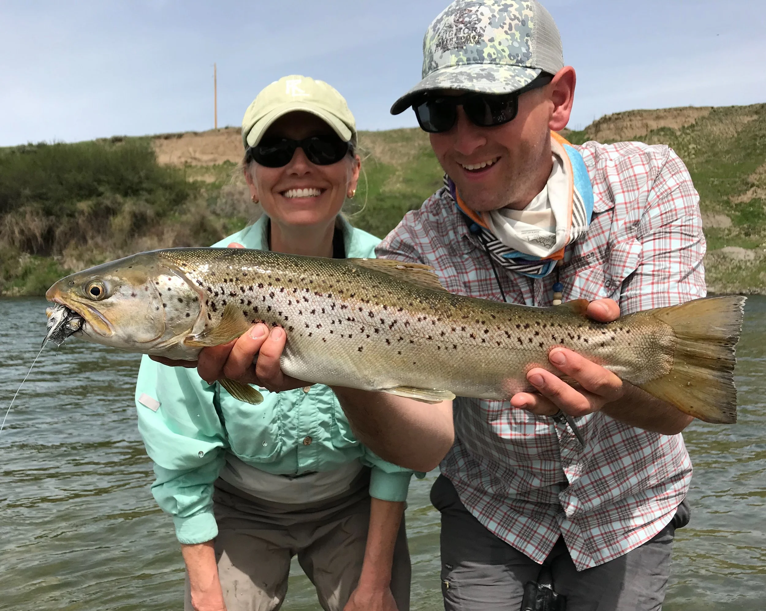 Two smiling people holding a large fish with a speckled body and a fishing hook in its mouth, standing in a river with grassy hills in the background.