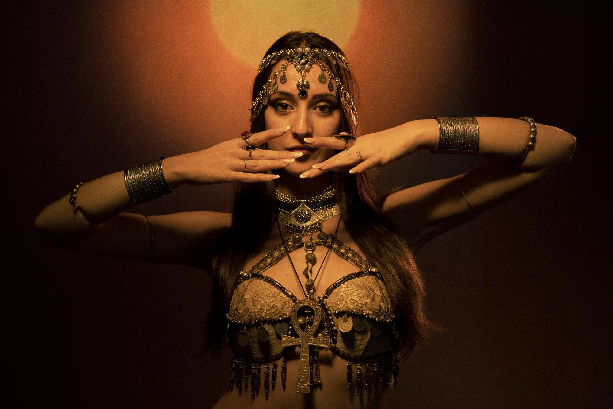 A woman dressed in Egyptian-style jewelry and accessories, posing with her hands framing her face, with warm lighting and a dark background.