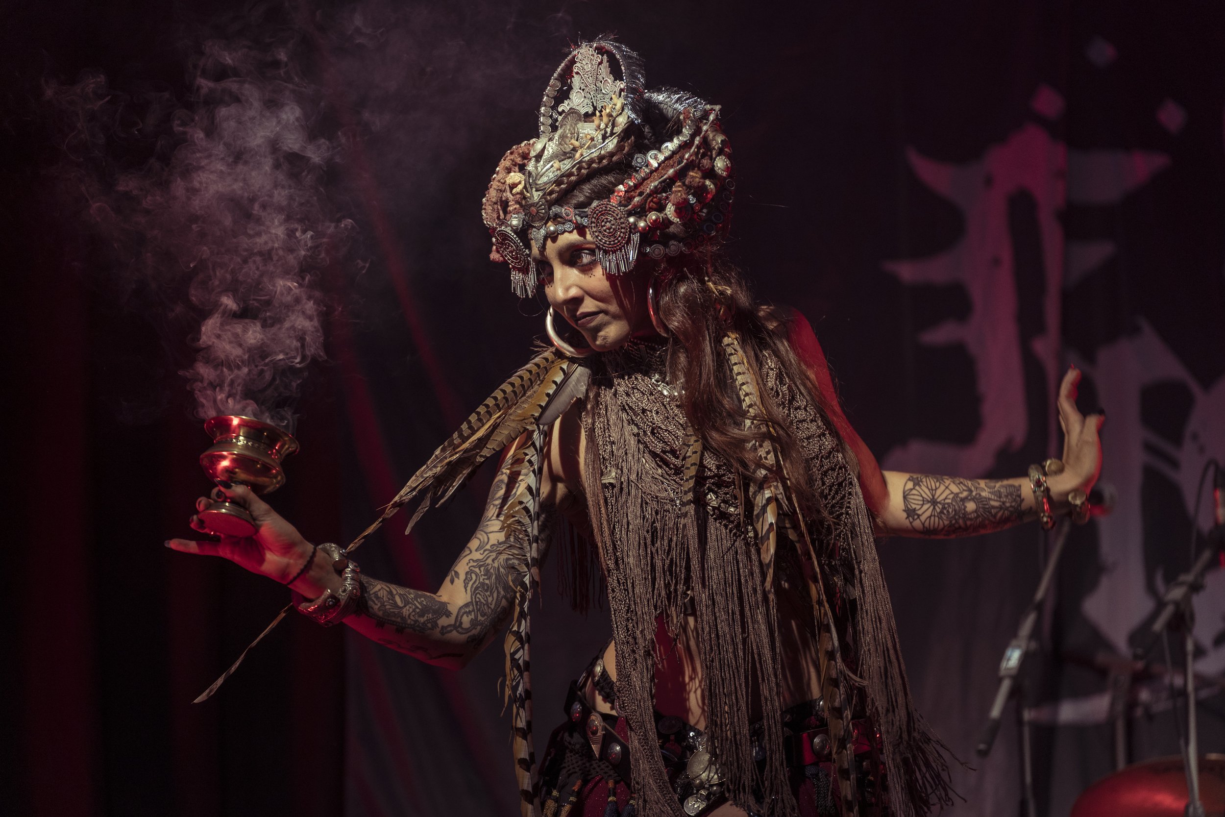A woman wearing an elaborate headdress and traditional attire, holding a smoking incense holder during a performance on stage.