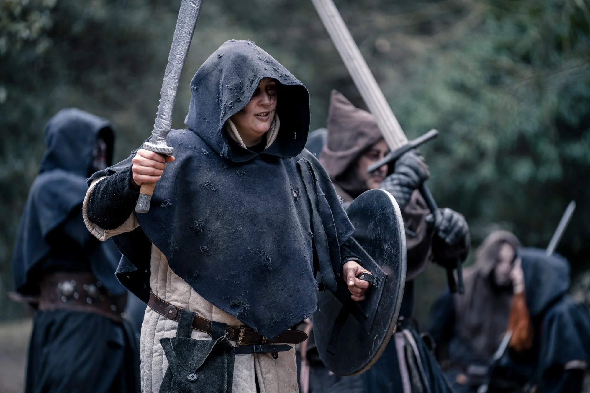 LARP fighter wearing a hood and holding a sword during a battle scene in a forest environment.