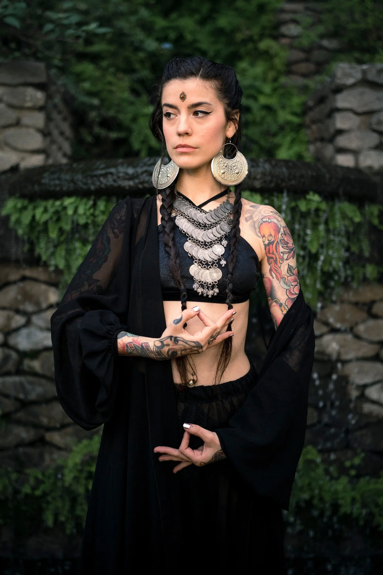 Portrait of tribal fusion dancer Sandra Roll wearing black attire and Indian-inspired jewelry, performing hand mudras in an urban garden in Barcelona.