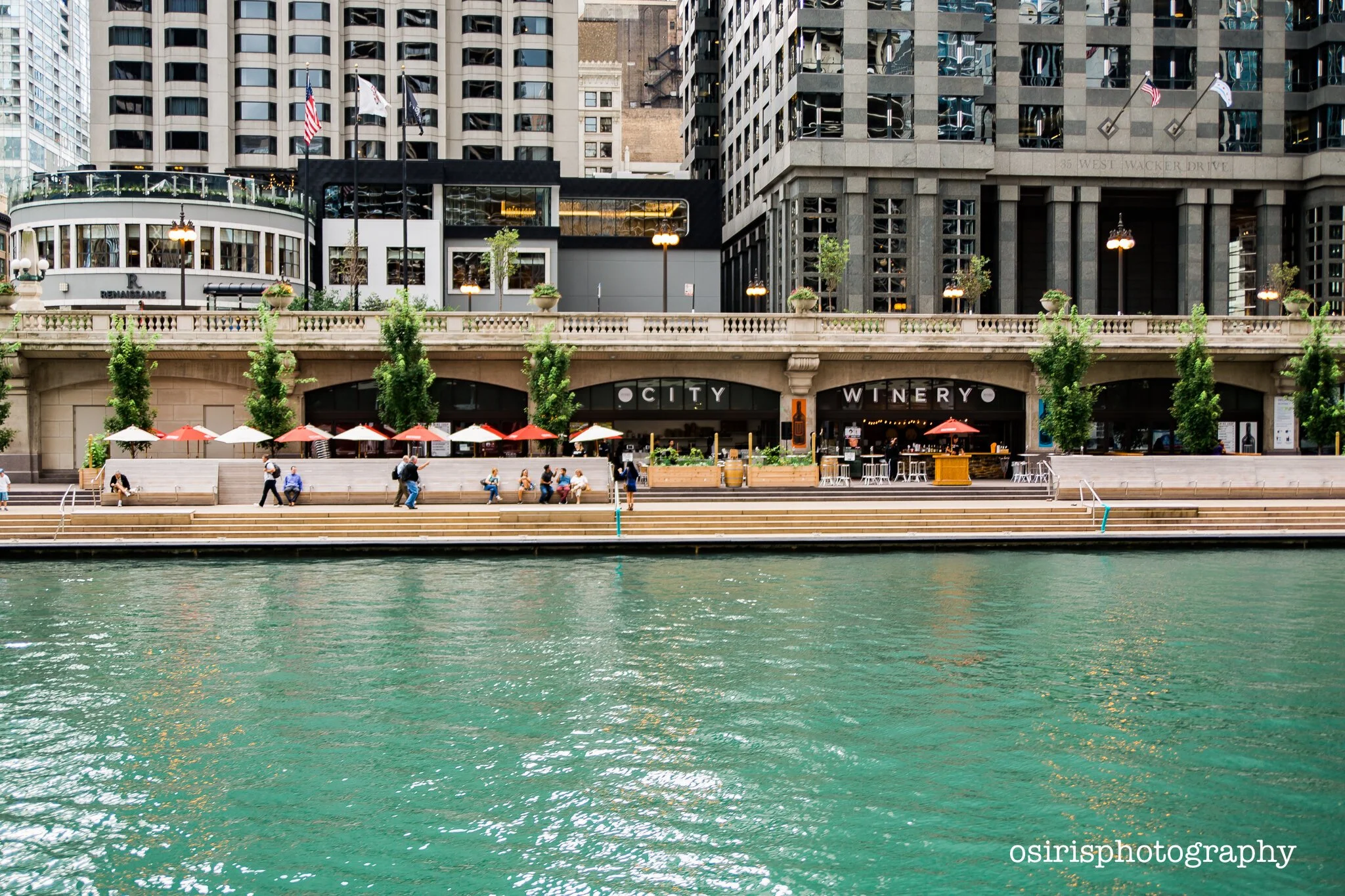People sitting and walking along a waterfront promenade in front of high-rise buildings, with a restaurant called 'CITY WINERY' and trees decorating the area.