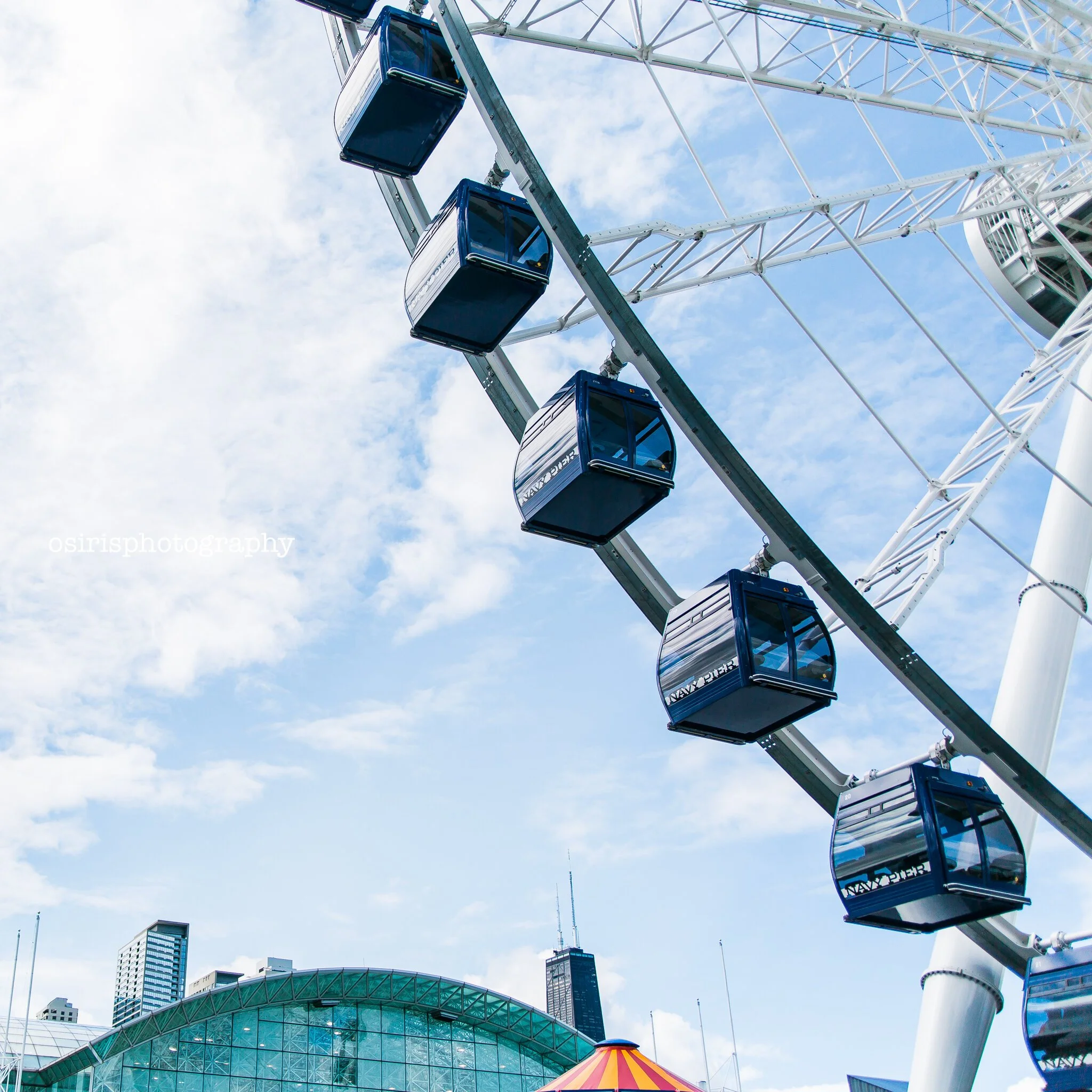 Close-up of a Ferris wheel with reflective blue gondolas against a blue sky with scattered clouds, in an outdoor amusement park setting.