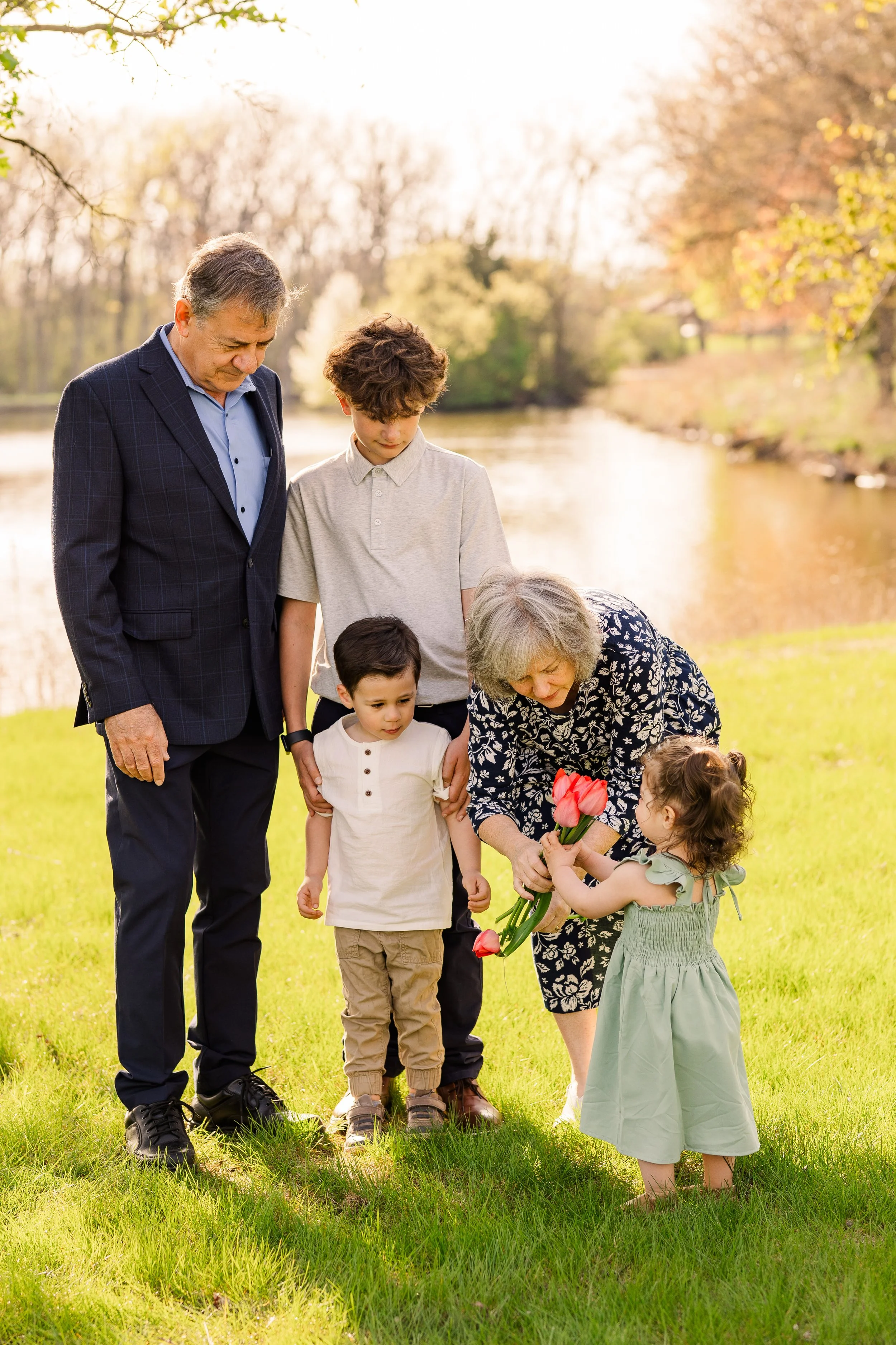 An elderly woman giving flowers to a young girl, with three boys and an older man standing nearby, outdoors by a river during spring.