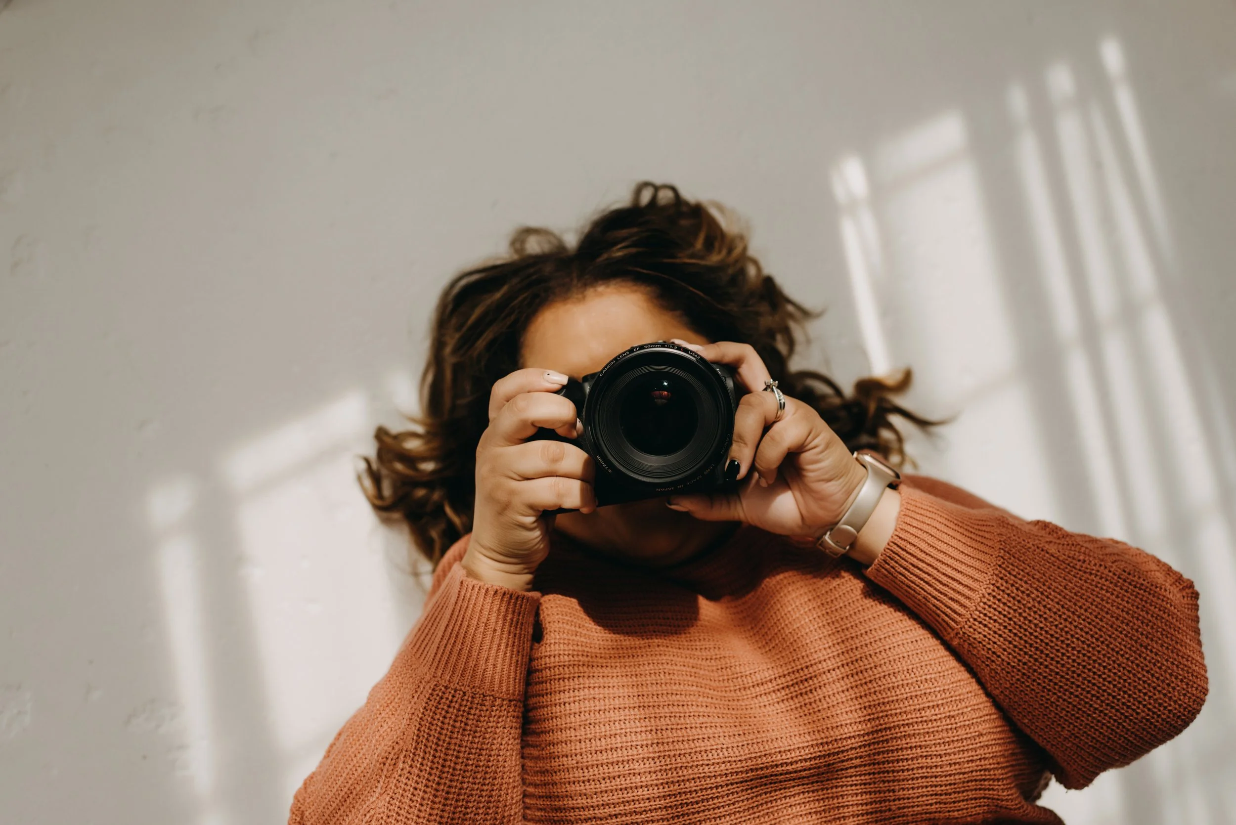 Person with curly hair taking a photo with a camera, wearing a rust-colored sweater, standing in front of a white wall with sunlight and shadows.
