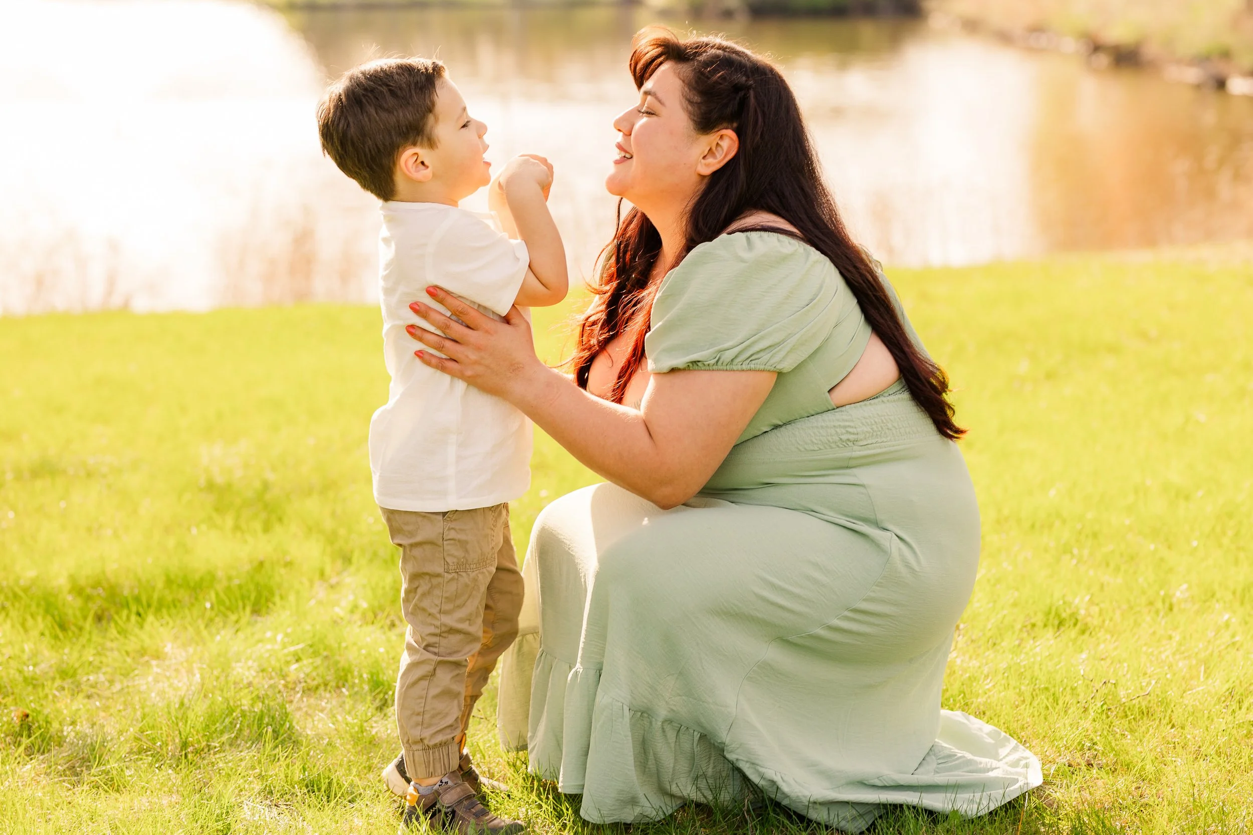 A woman in a light green dress kneels on the grass, holding and smiling at a young boy by a lake on a sunny day.