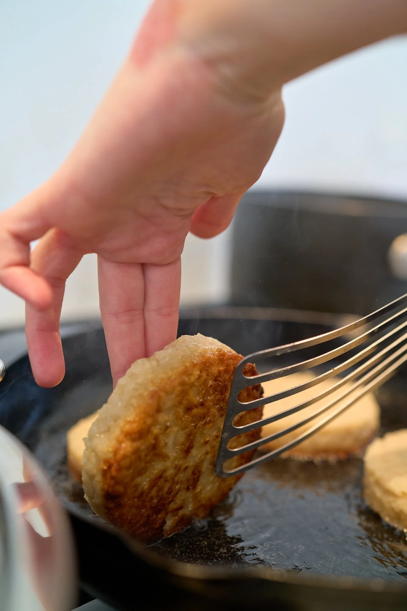 A close-up of a hand holding a fork with a golden-brown, breaded food item over a frying pan on the stove.