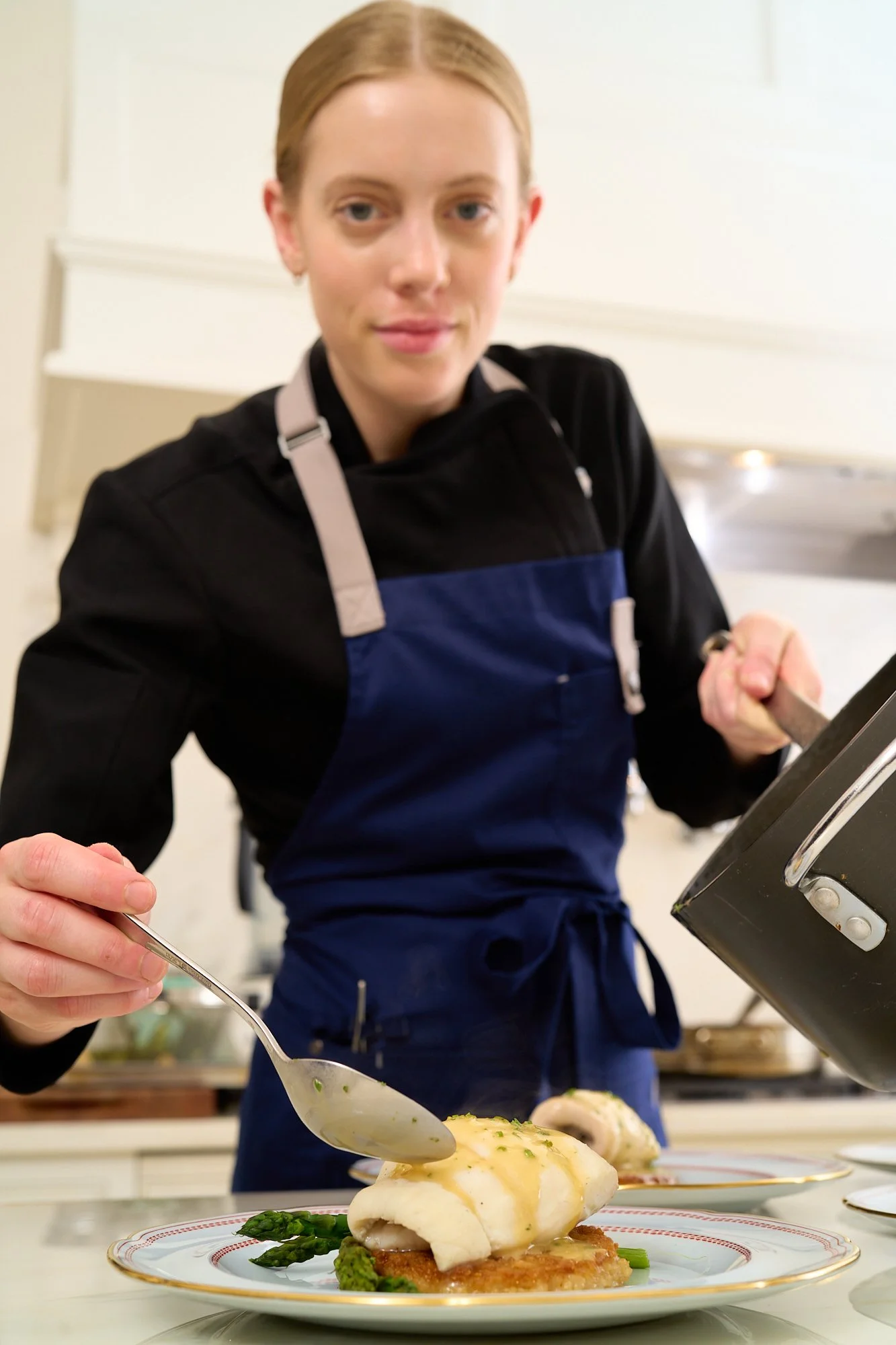 A woman in a black shirt and blue apron garnishes a plate of food, which includes fish, asparagus, and sauce, in a kitchen setting.