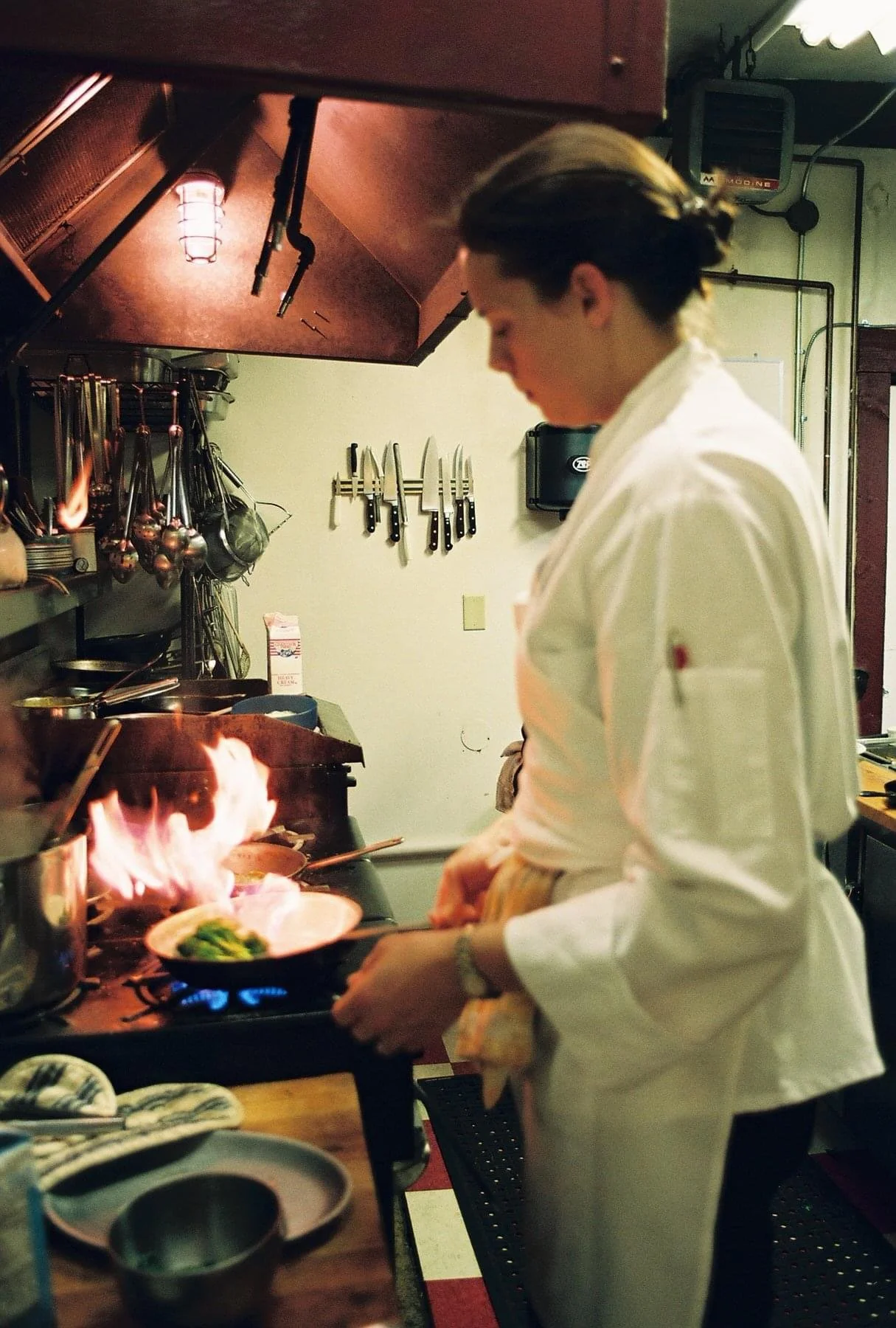 Chef cooking on a stove with flames, wearing a white uniform in a professional kitchen.