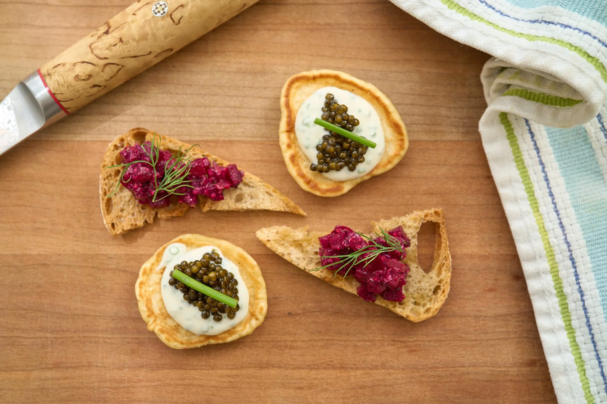 Four pieces of canapés on a wooden surface, with two topped with beetroot and garnished with dill, and two with a dollop of cream, capers, and a green garnish, sitting near a cloth and a rolling pin.