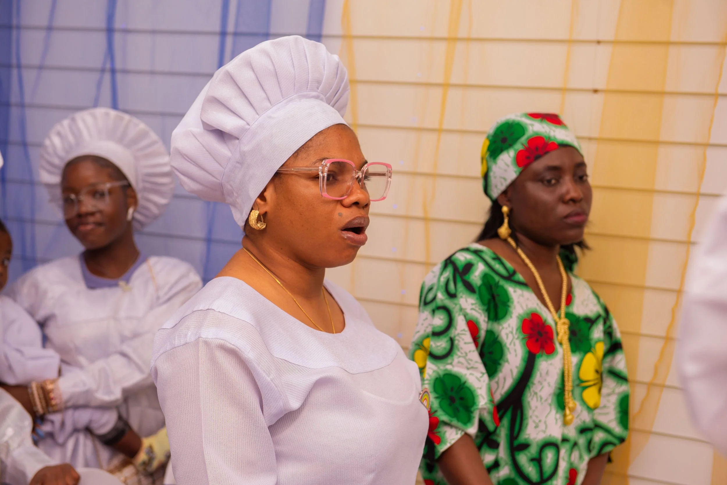 Women dressed in traditional and cultural attire, standing in a room decorated with yellow and blue drapery.