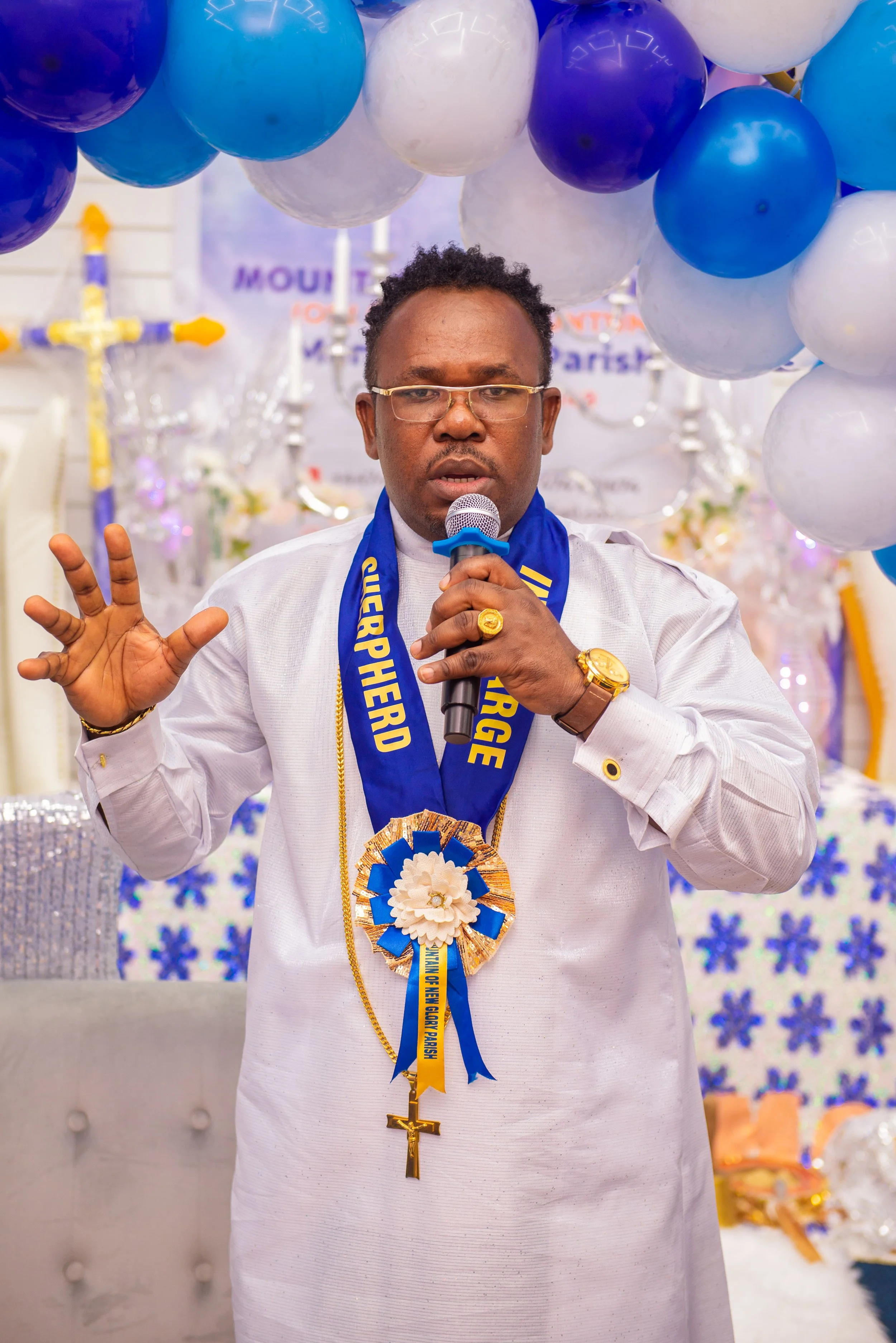 A man wearing glasses and a white traditional outfit, speaking into a microphone at an event decorated with blue, white, and purple balloons and party decorations.