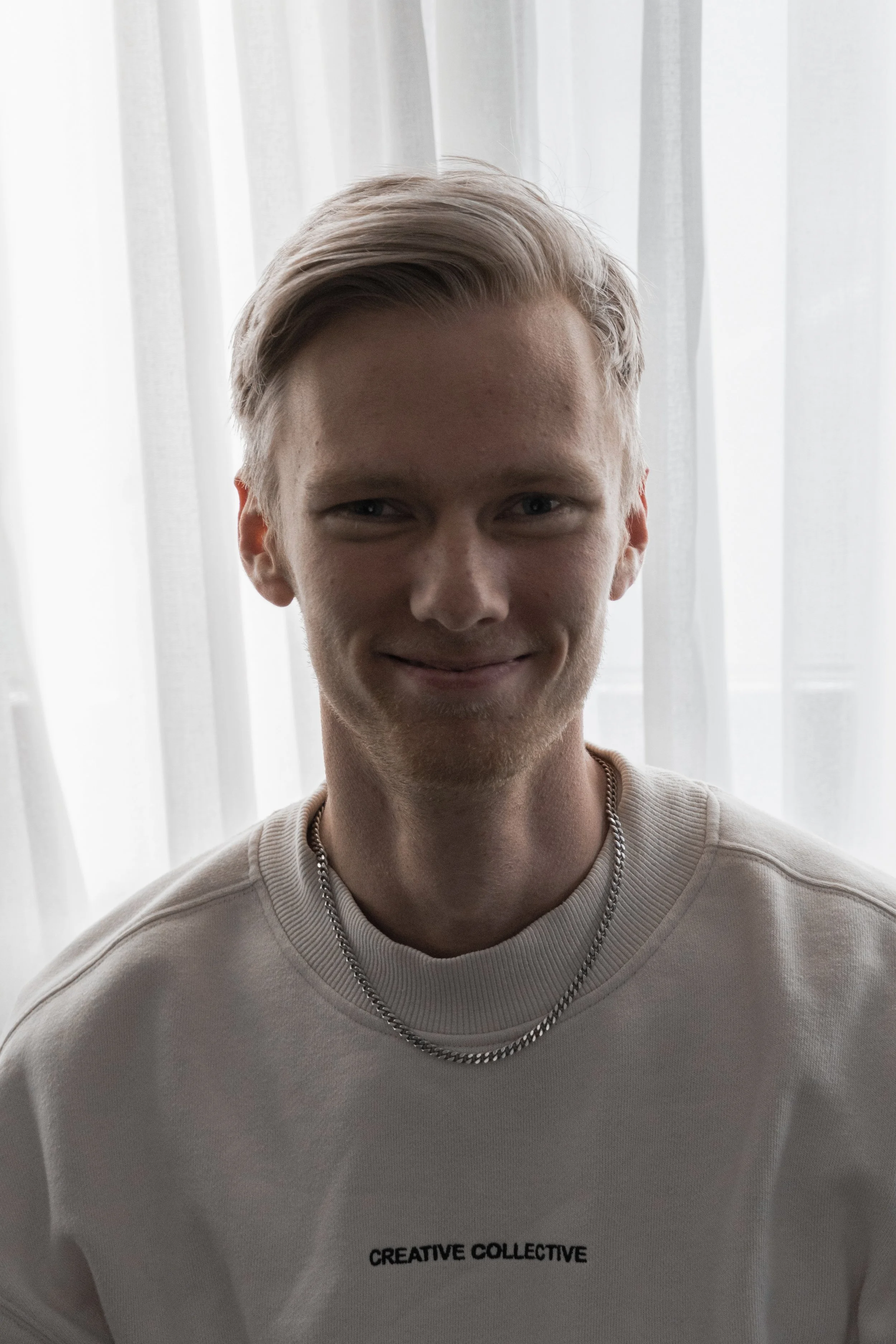 Young man with blonde hair smiling, wearing a light-colored sweatshirt with 'CREATIVE COLLECTIVE' written on it, standing in front of white curtains.