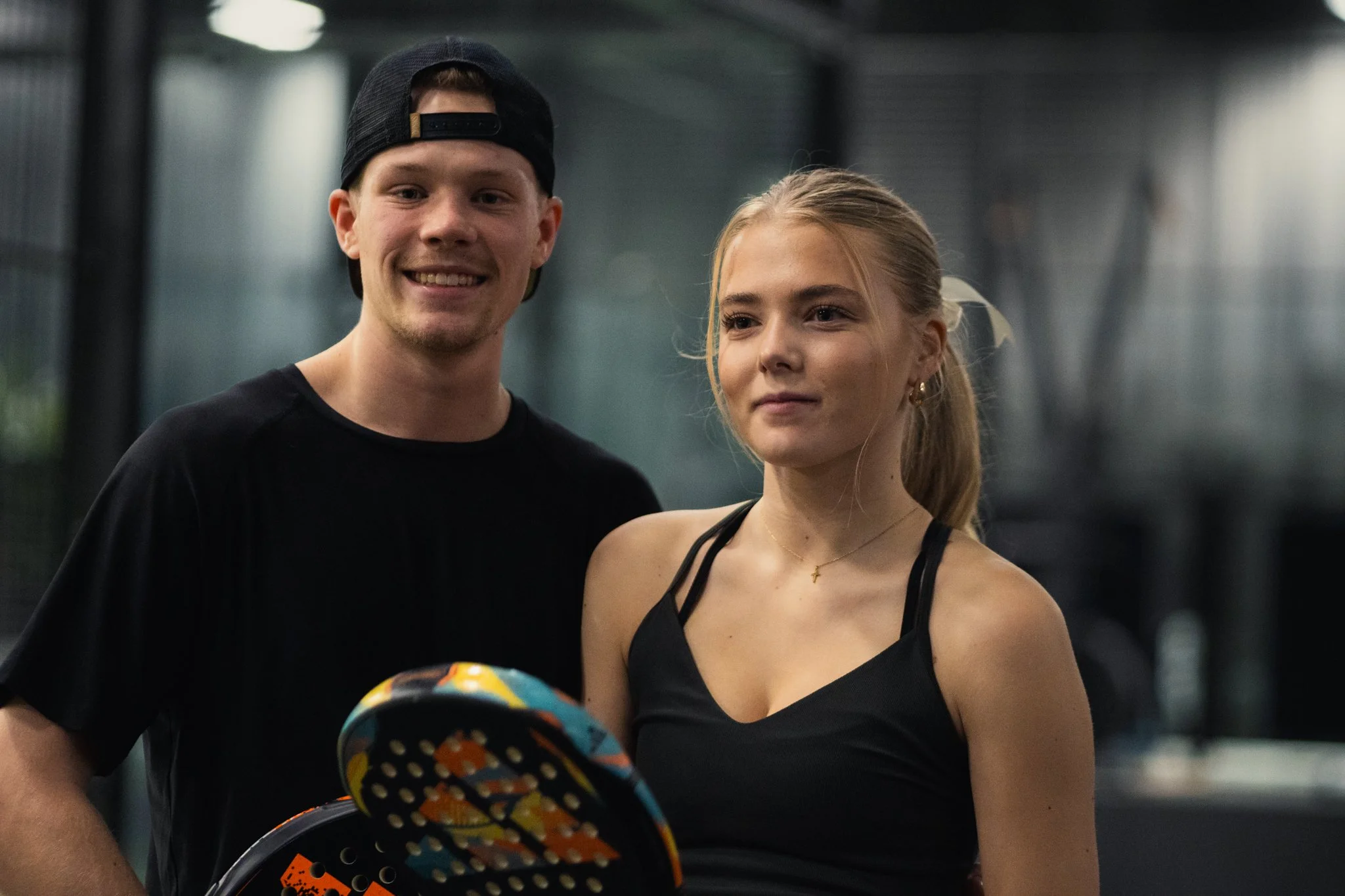 A young man and woman stand together indoors, with the man smiling and the woman looking serious, holding a colorful paddle tennis racket.