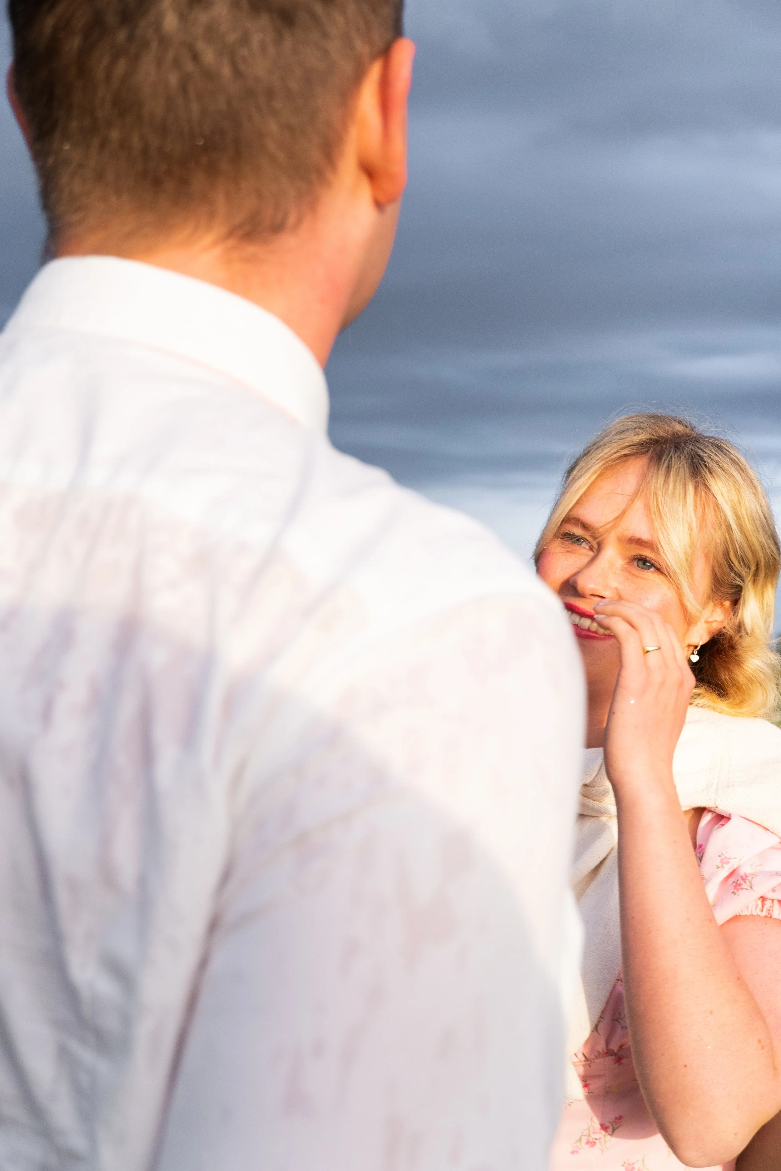 A woman is smiling and covering her mouth with her hand while looking at a man, on a cloudy day outdoors.