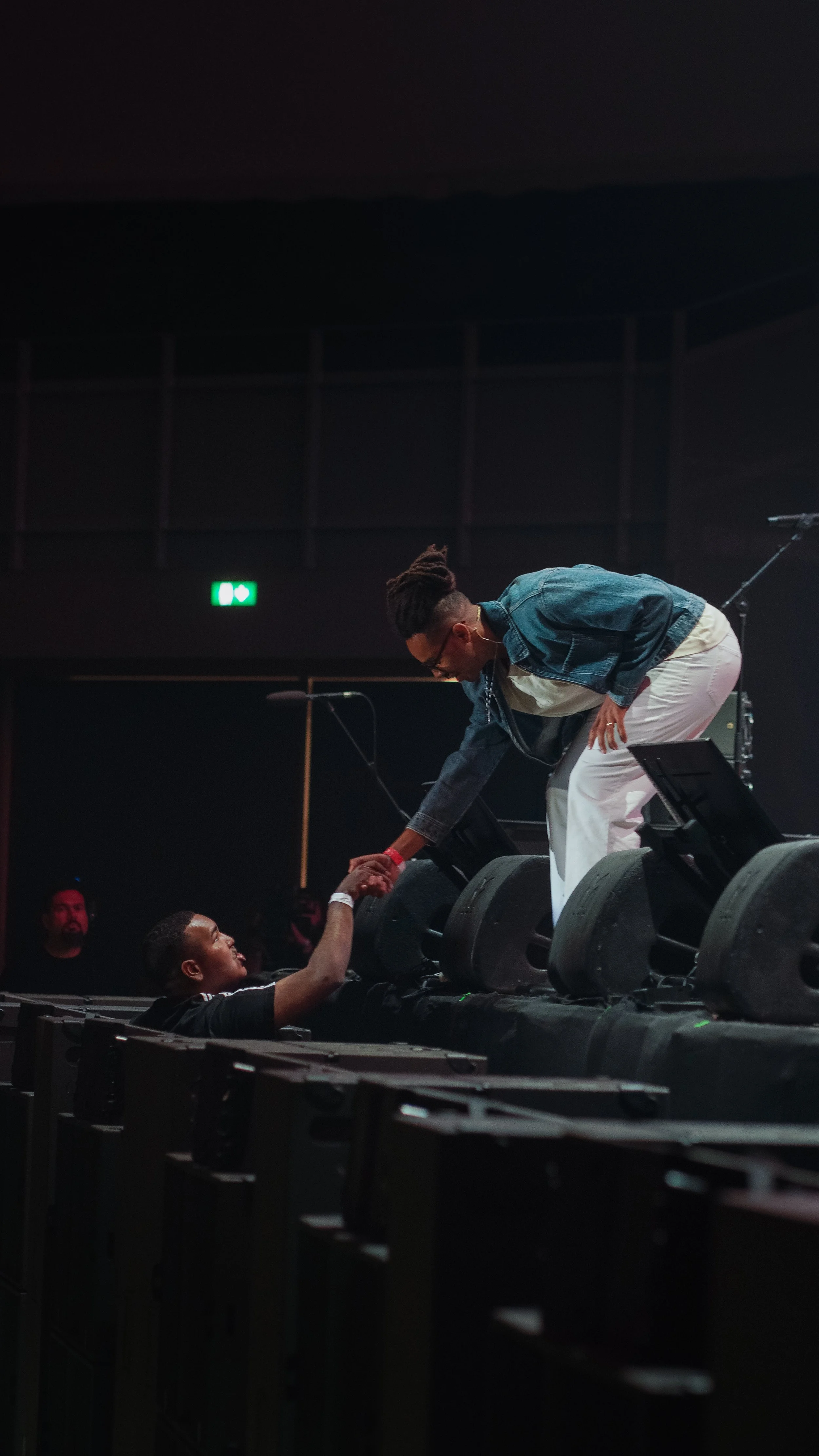 A man on stage leaning down to shake hands with a concert attendee who is lying on the stage, in a dark indoor concert venue.