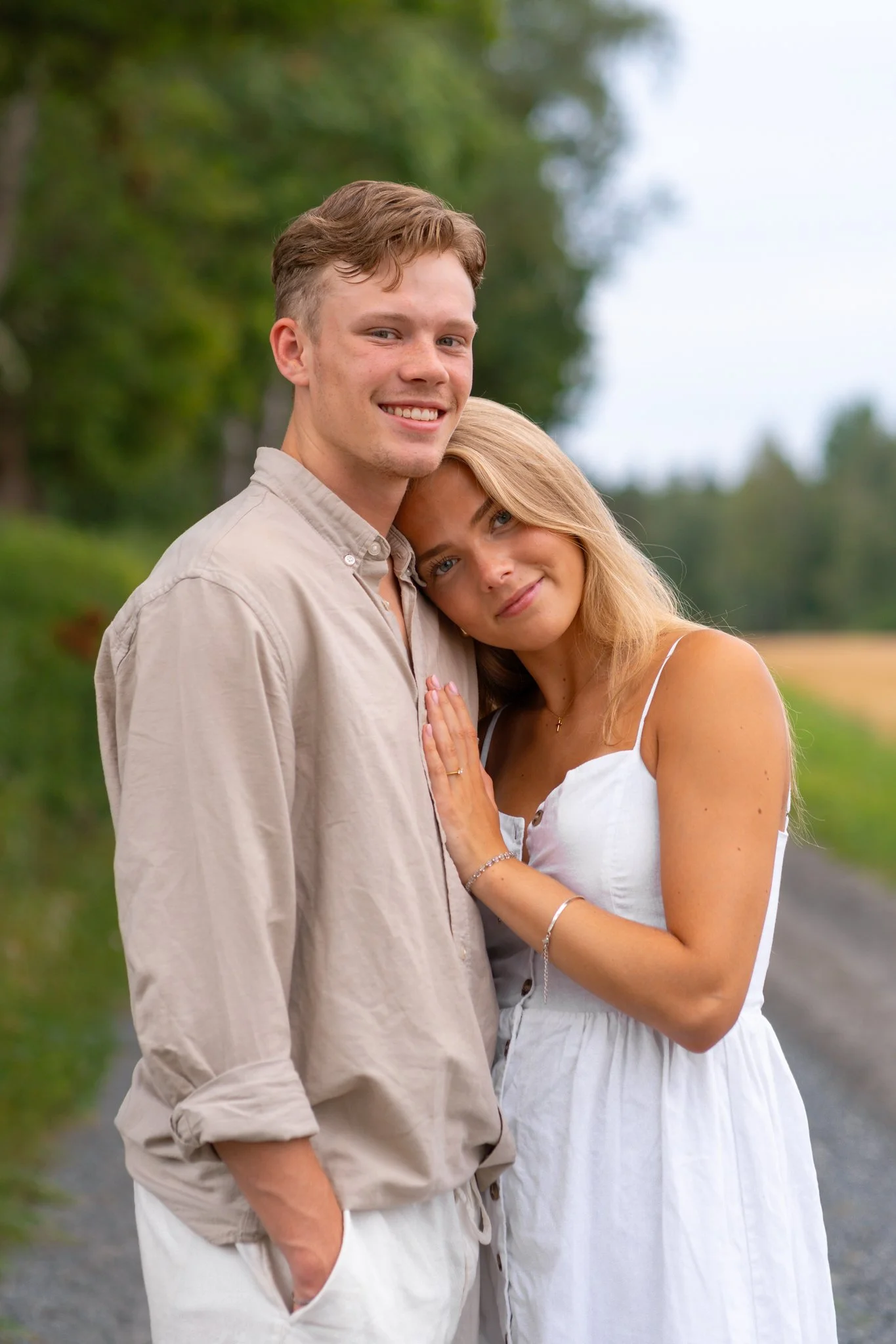 A couple standing on a rural road outdoors, with tall trees in the background. The man is smiling at the camera while the womanrests her head on his shoulder, with her hand on his chest, both dressed in light-colored casual clothing.