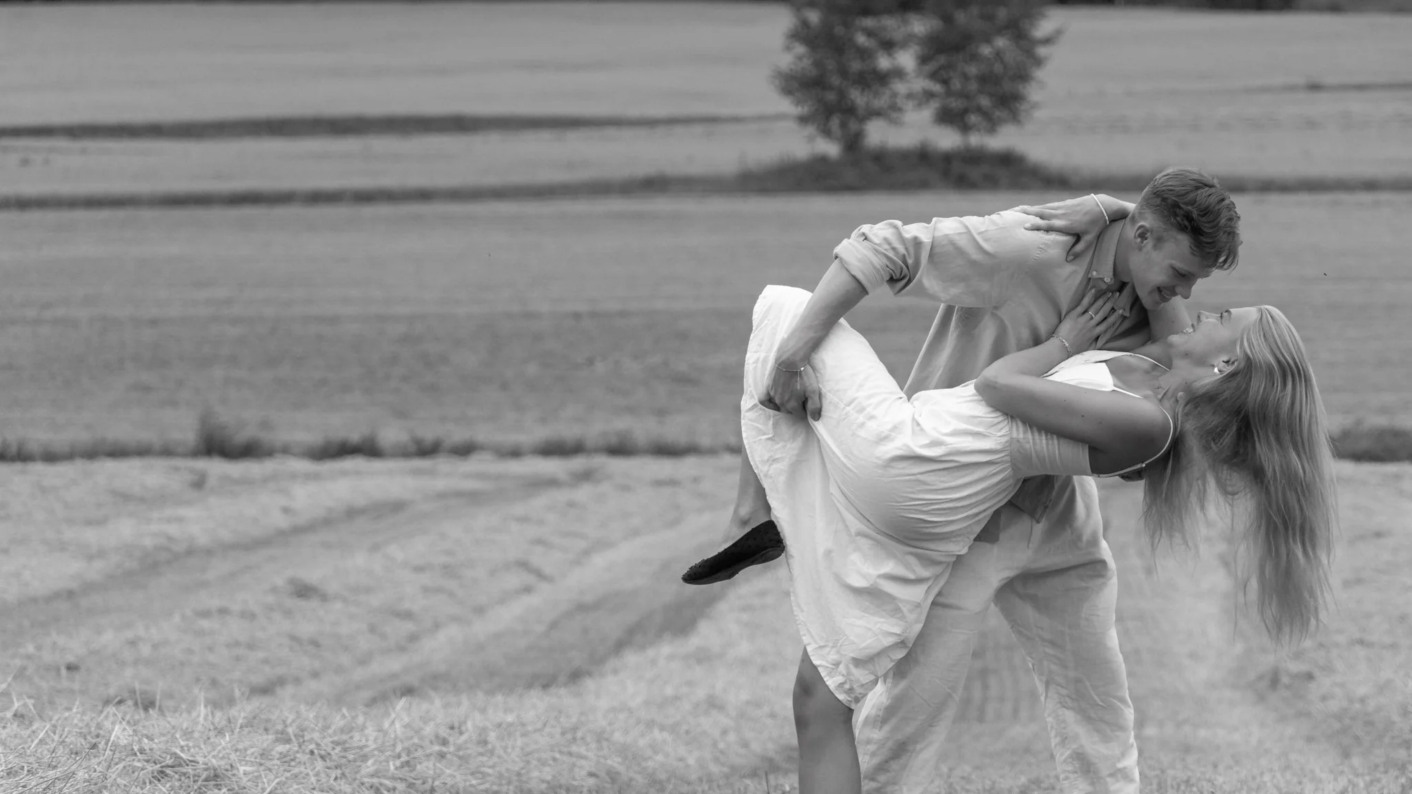 A smiling couple is outdoors on a grassy field, with the man holding the woman in a dip motion, both enjoying each other's company.