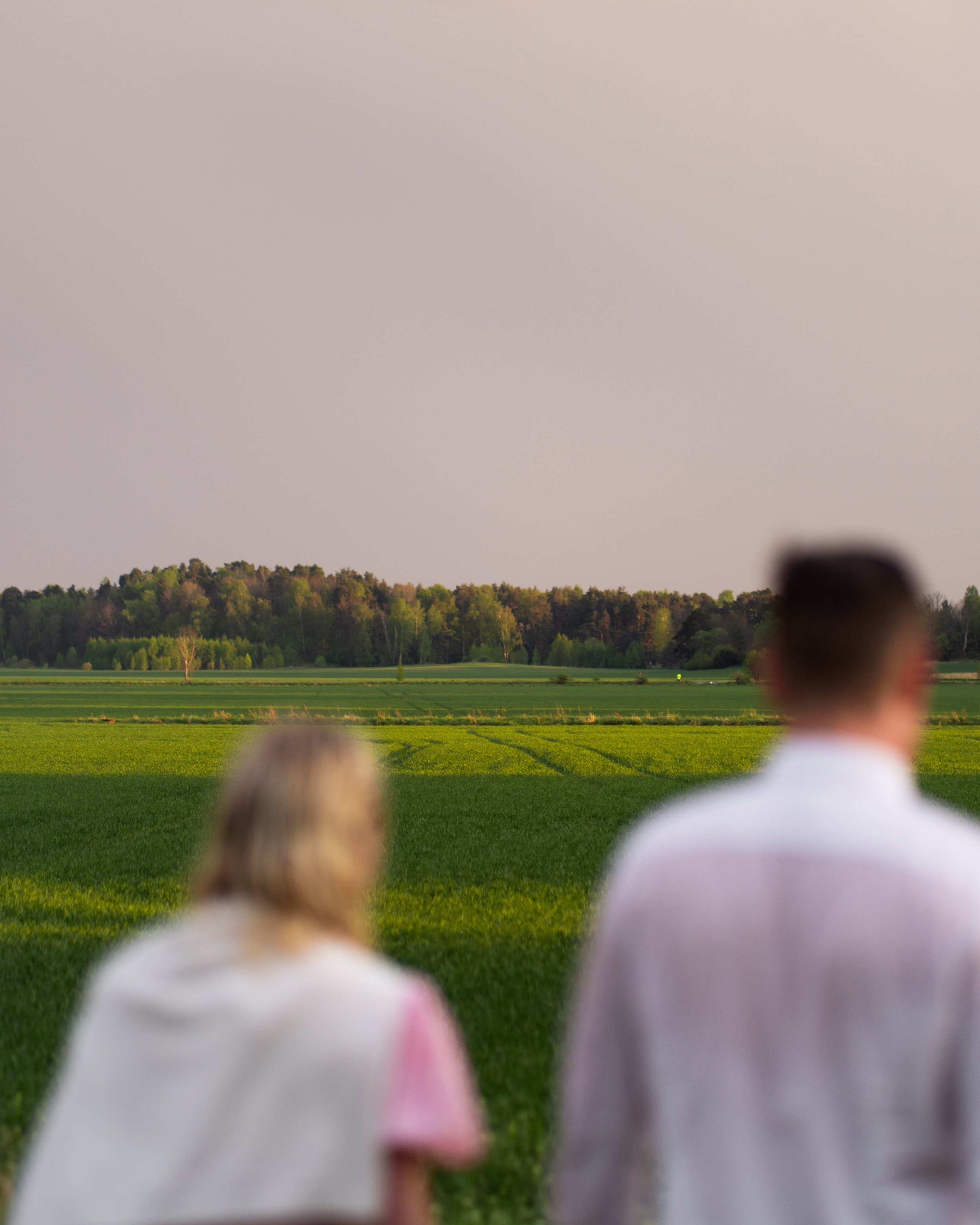 Blurry image of two people sitting on a grassy field, facing a distant tree line and a clear sky.