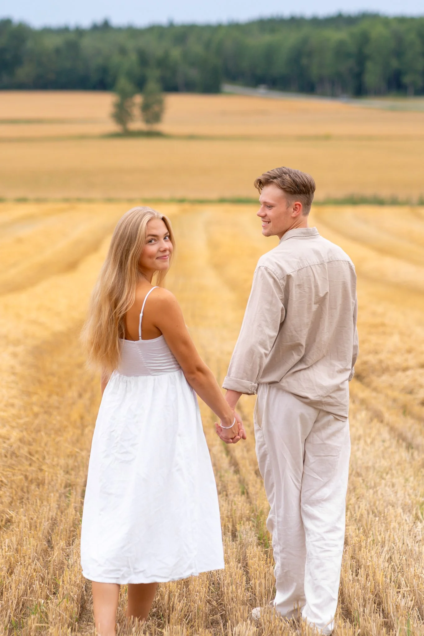 A couple holding hands in a wheat field, with a backdrop of trees and a blue sky.
