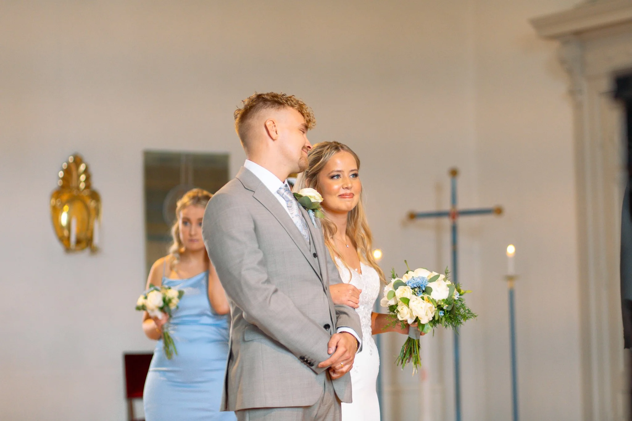 A couple getting married in a church, with a woman in a blue dress holding a bouquet of white flowers in the background.