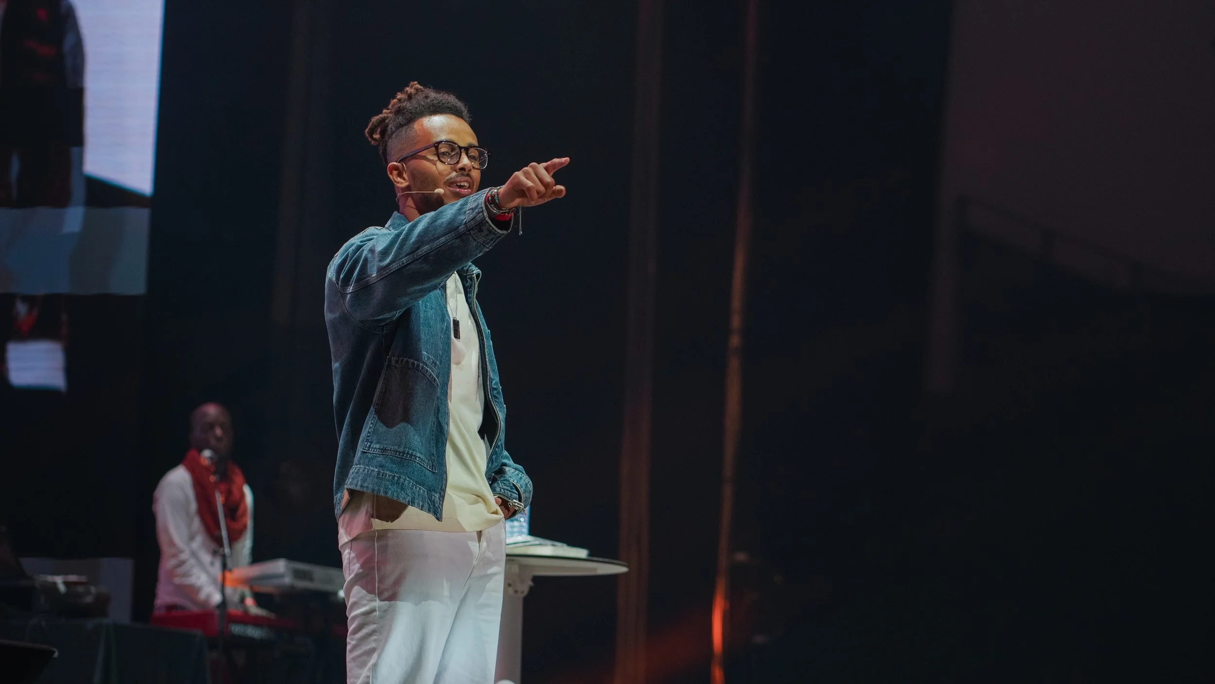 A man with glasses and a microphone headset speaking on stage, pointing forward, with a person in the background playing keyboard.