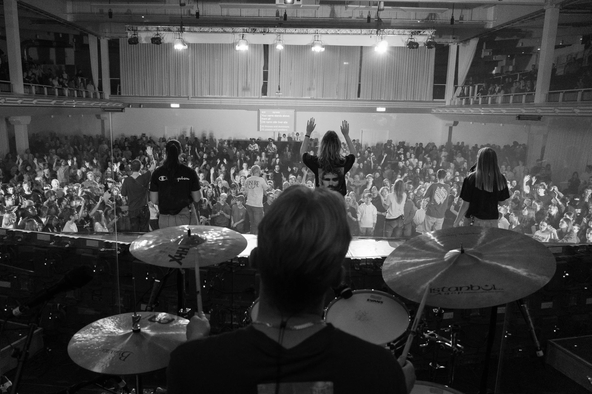 A black and white photo of a live concert from behind the drum set, showing a crowd on the stage, with fans raising their hands in the air, in a large indoor venue.