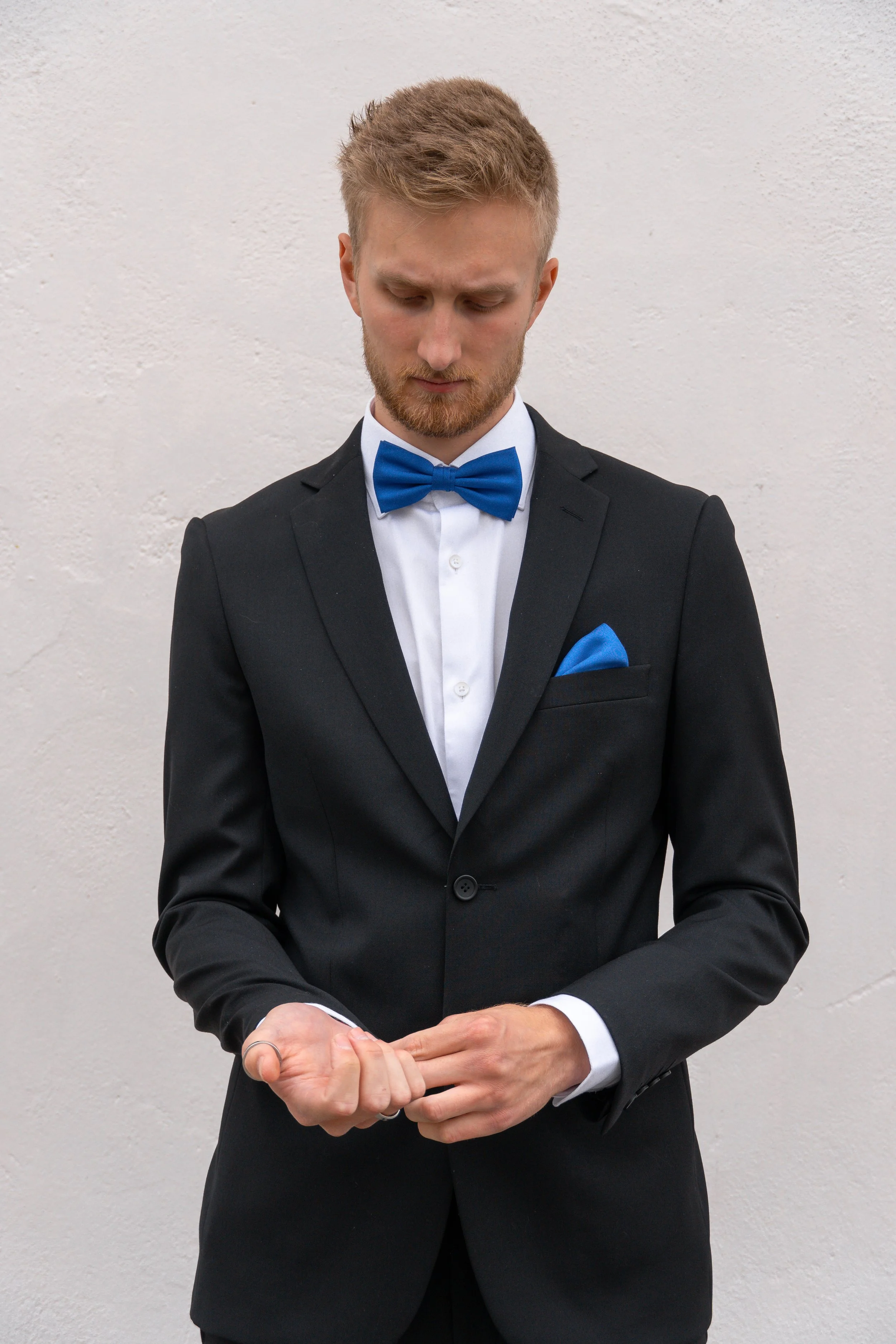 A man in a black tuxedo with a white shirt, blue bow tie, and blue pocket square adjusts his cuff against a plain white wall.