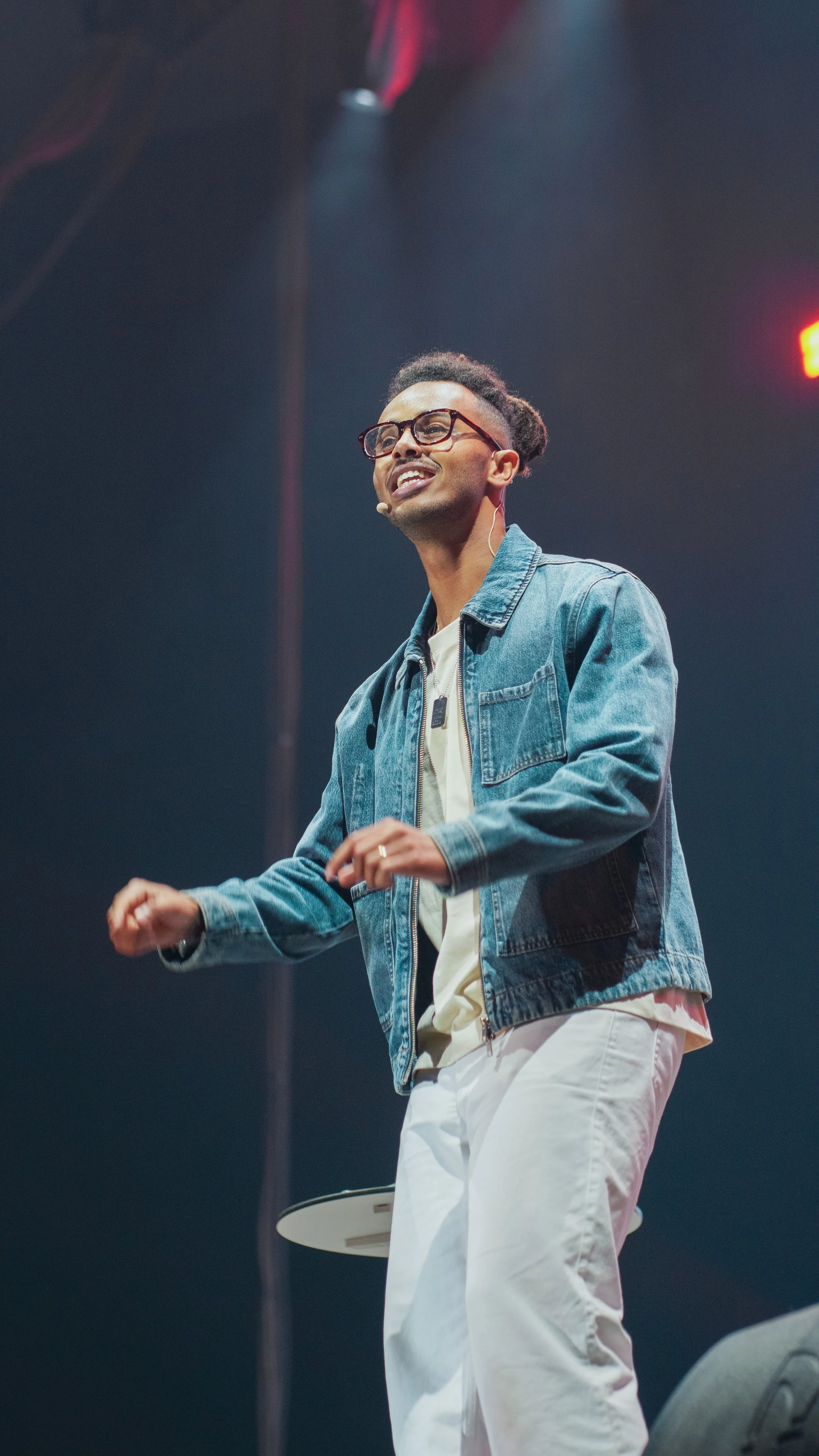 A man wearing glasses and a denim jacket smiling and speaking on stage with a microphone headset.