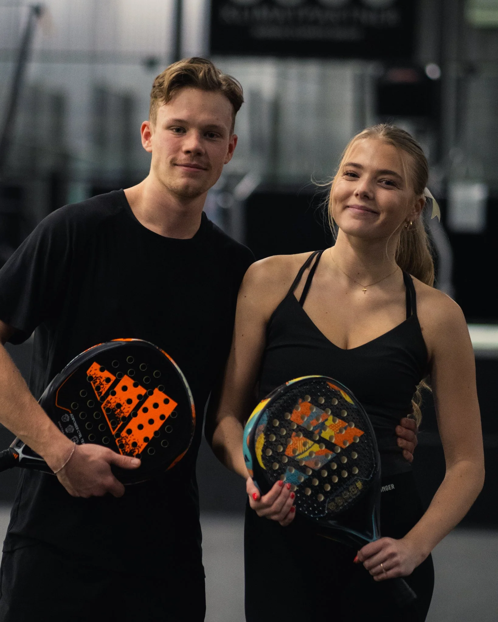 A young man and woman holding padel rackets in an indoor sports facility, smiling at the camera.