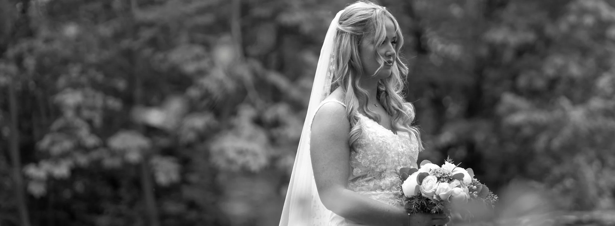 Black and white photo of a bride holding a bouquet of flowers, standing outdoors with blurred trees in the background.