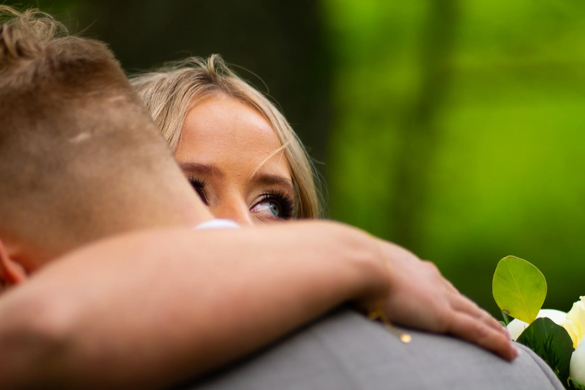 A woman with blonde hair and makeup resting her head on a man's shoulder outdoors, with green foliage in the background.