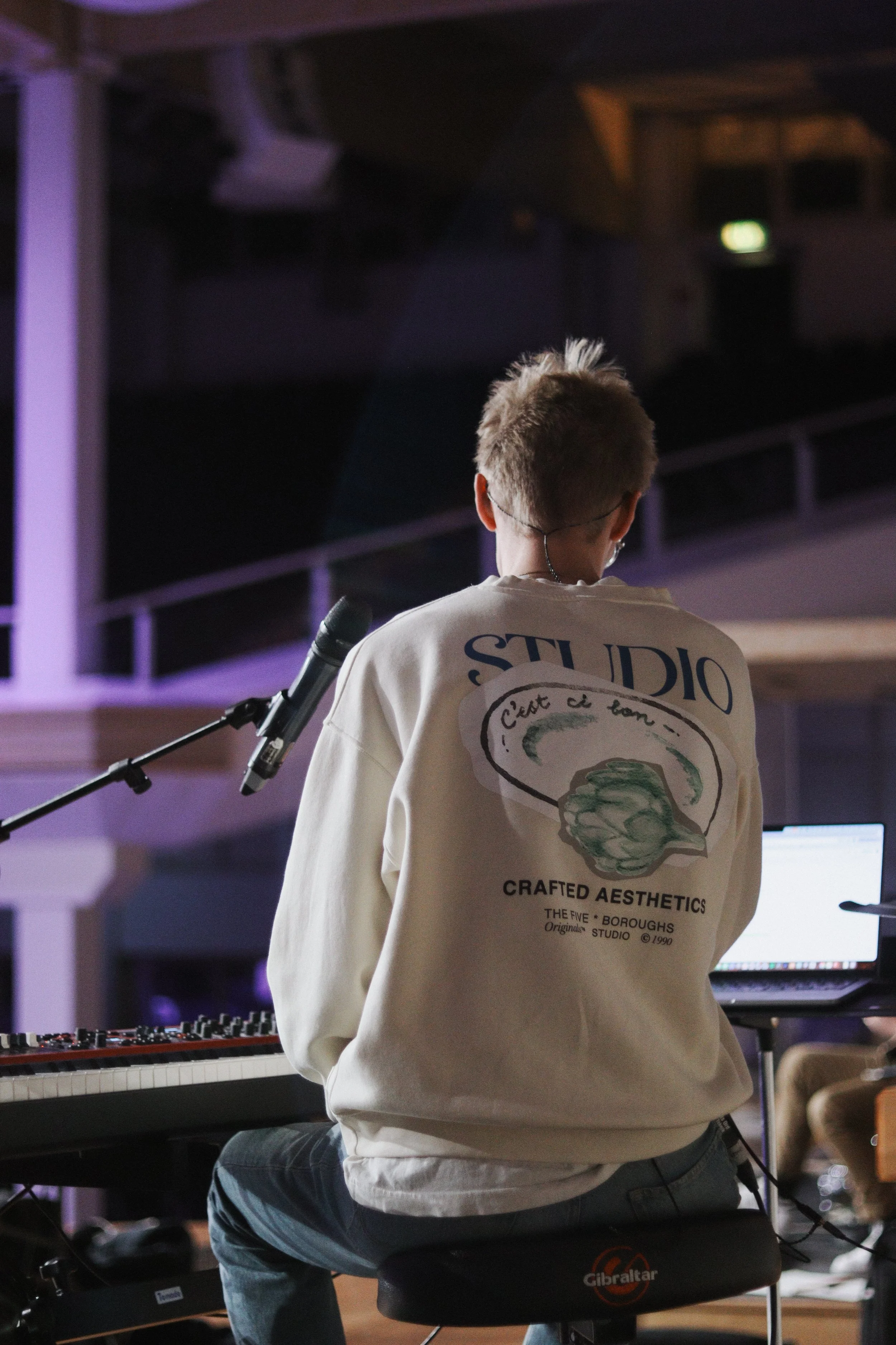 Back of a pianist in a white hoodie sitting at a keyboard and a laptop, facing away, in a dimly lit indoor church space.