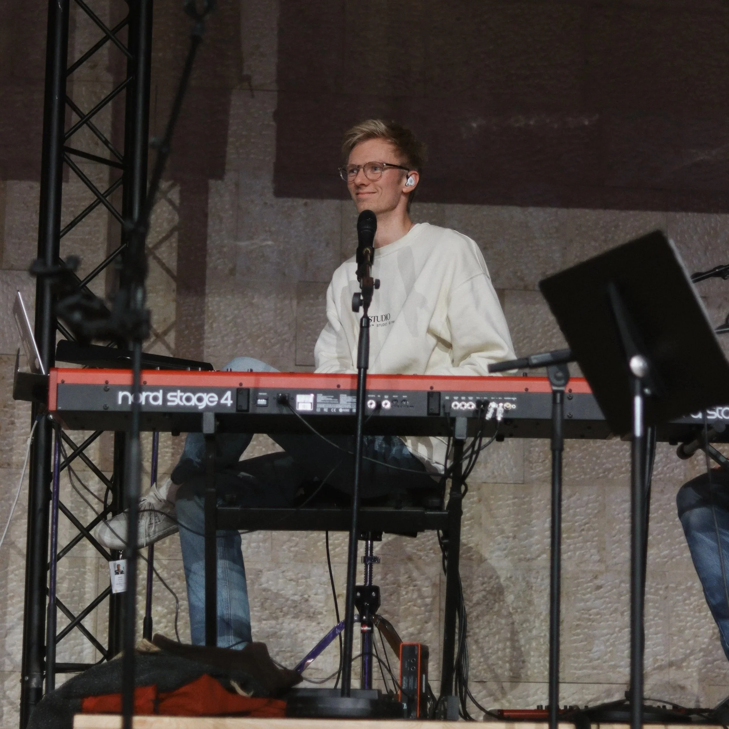 A young man playing a Nord Stage 4 keyboard on stage in Uppsala, with a microphone in front of him, smiling and wearing glasses and a white sweatshirt.