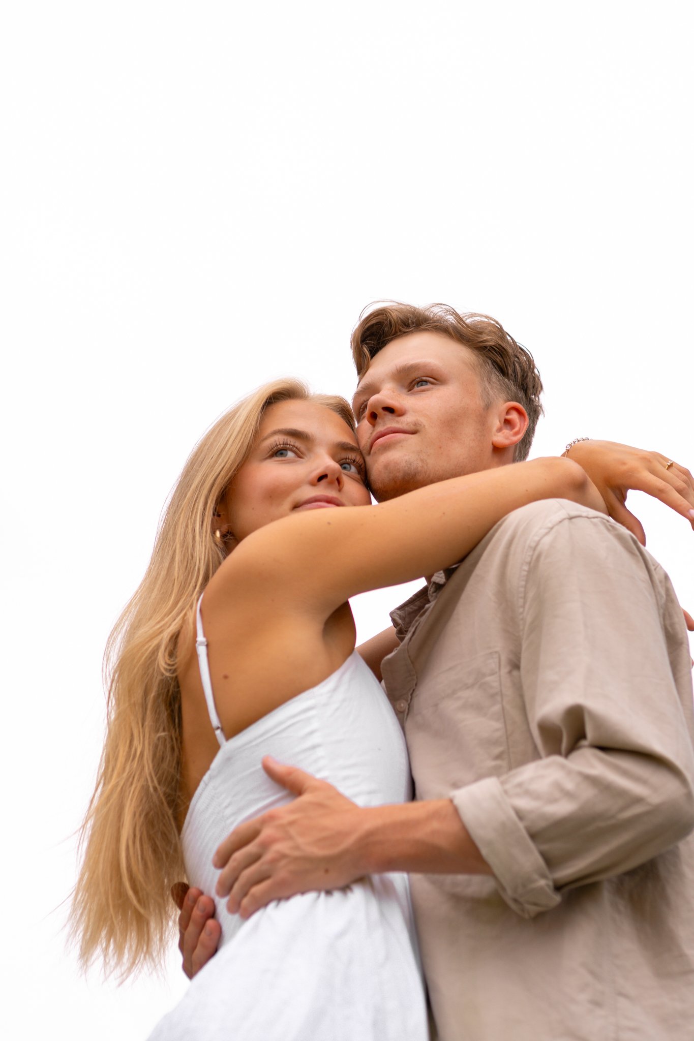 A young man and woman embrace outdoors against a white sky, gazing into each other's eyes with affection.