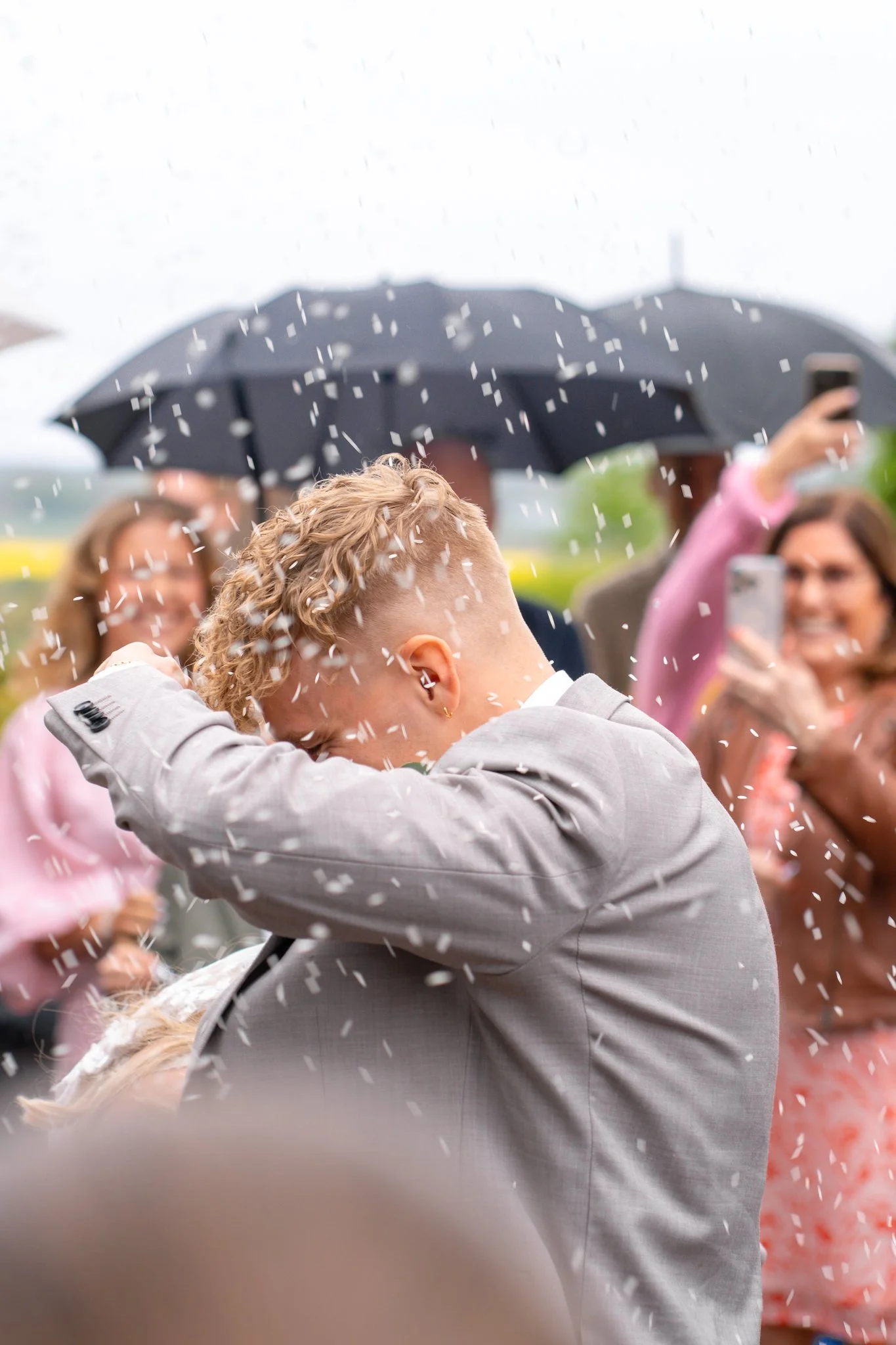 Person with curly blonde hair in a light-colored suit celebrating with confetti at a wedding or celebration, with guests taking photos and a person holding an umbrella in the background on a rainy day.