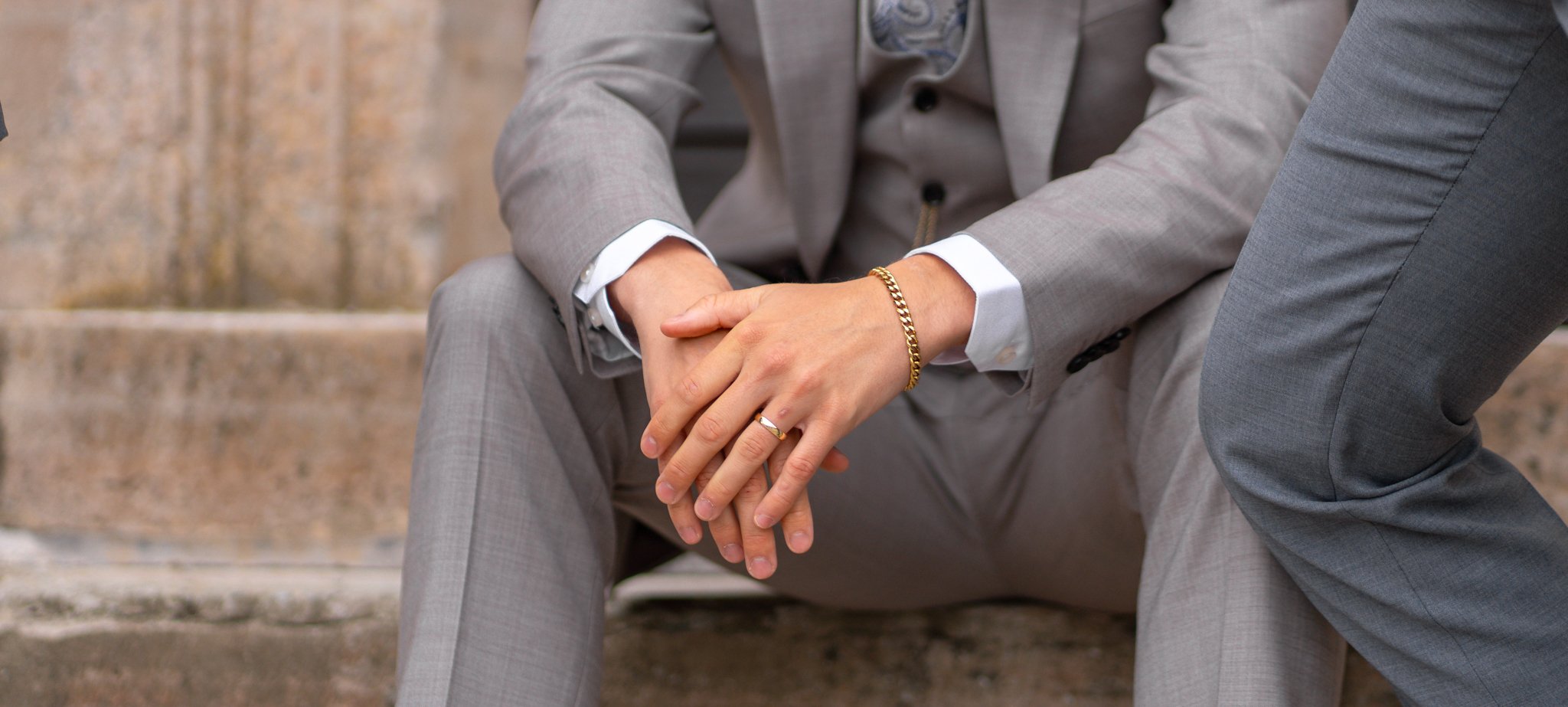 Person in a light gray suit sitting on stairs, hands resting on knees, wearing a wedding band and gold bracelet.