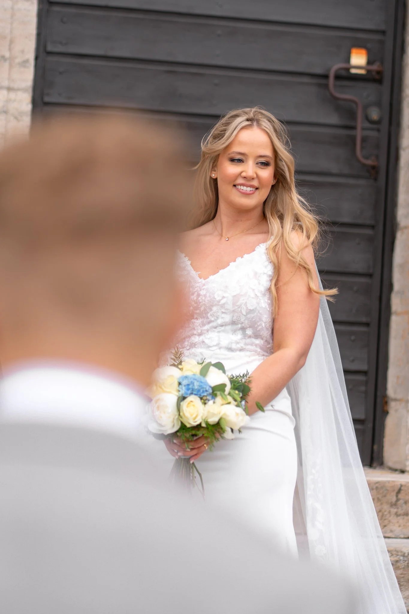 A bride in a white wedding dress holding a bouquet of yellow, white, and blue flowers, smiling.
